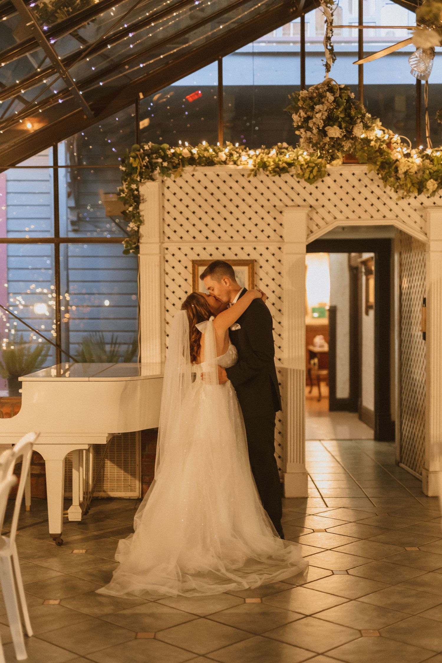 Couple embraces under white lattice arch with twinkling lights and greenery in romantic indoor wedding venue.