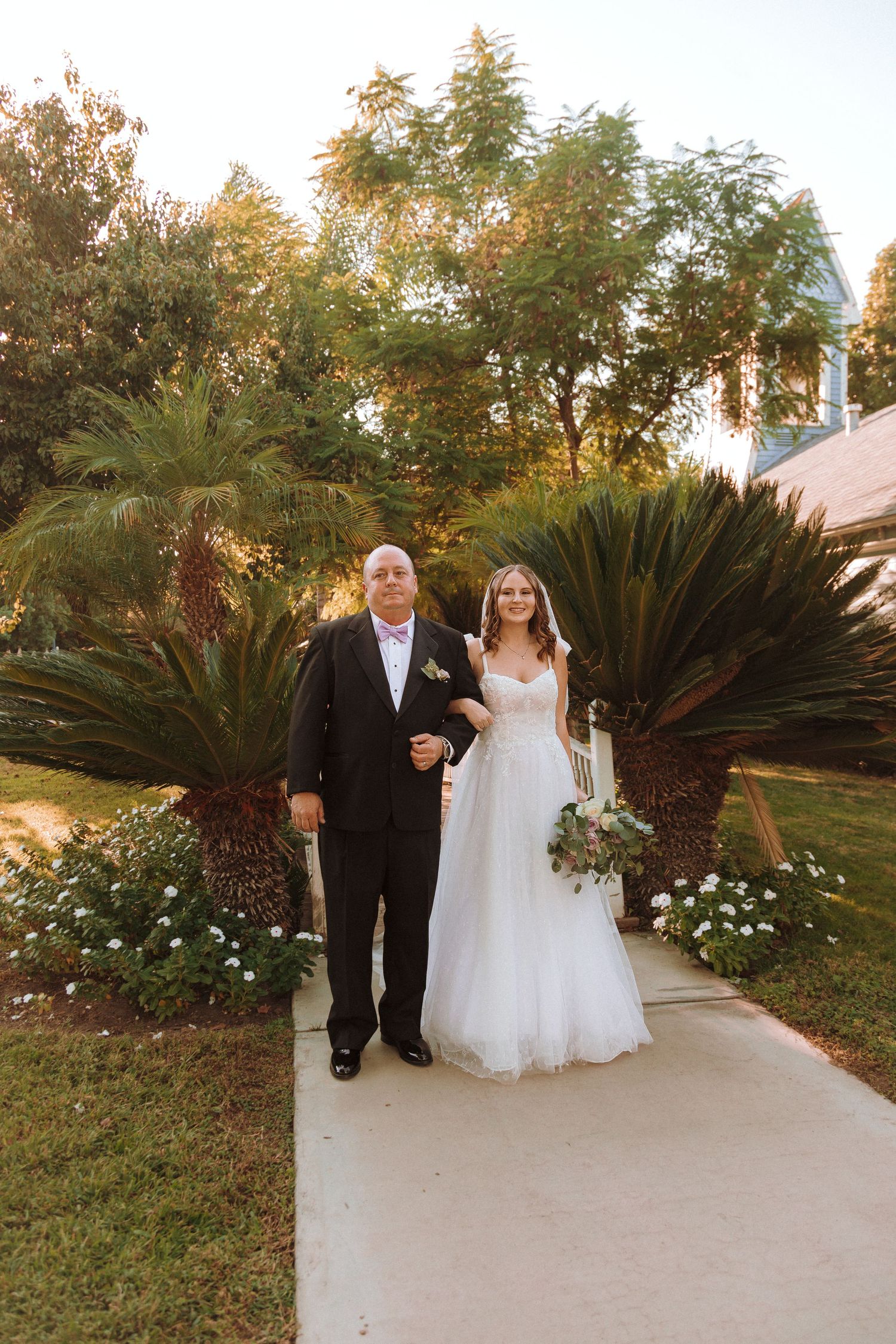 Wedding couple poses on garden pathway surrounded by tropical plants and flowers.