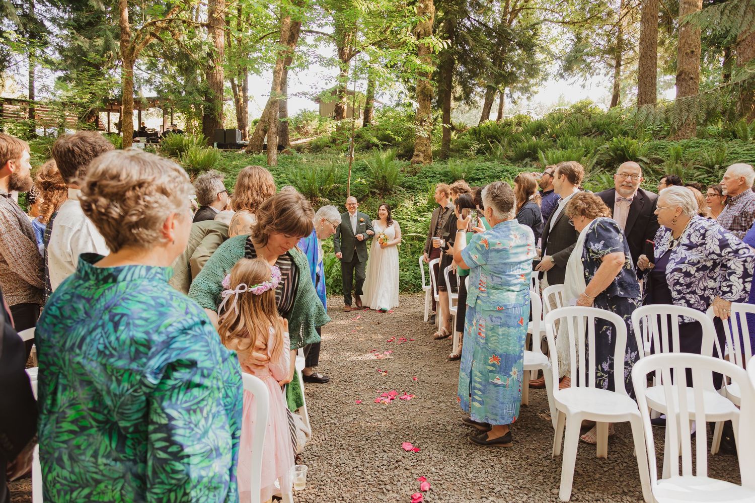 Wedding guests stand along a path decorated with rose petals in a forest setting.