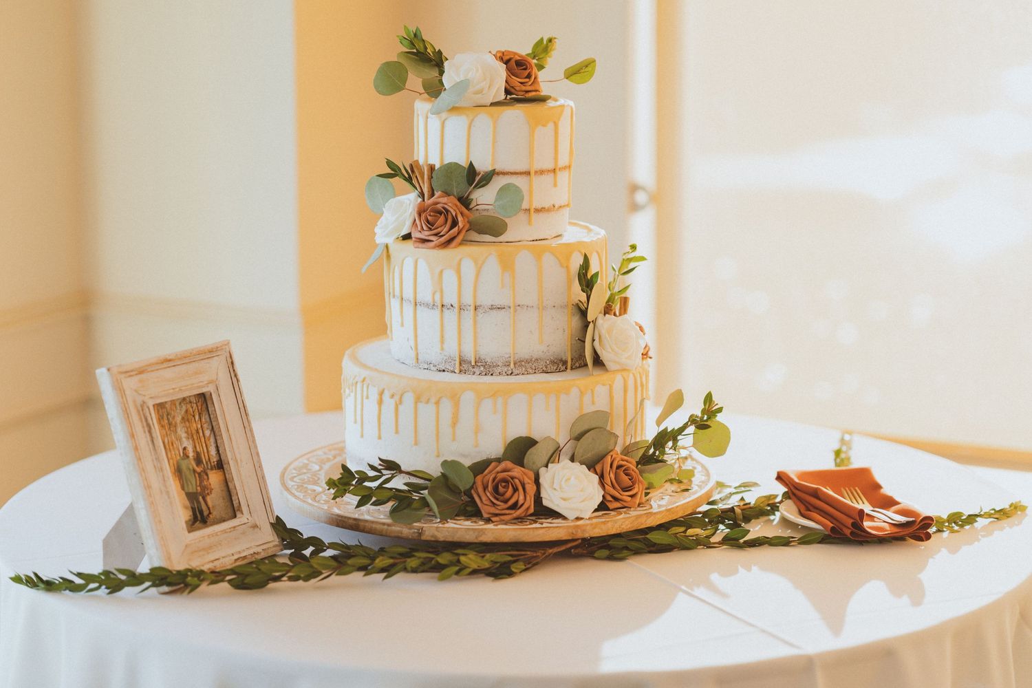 Wedding cake display with gold drip detail, floral decorations and greenery on white table.