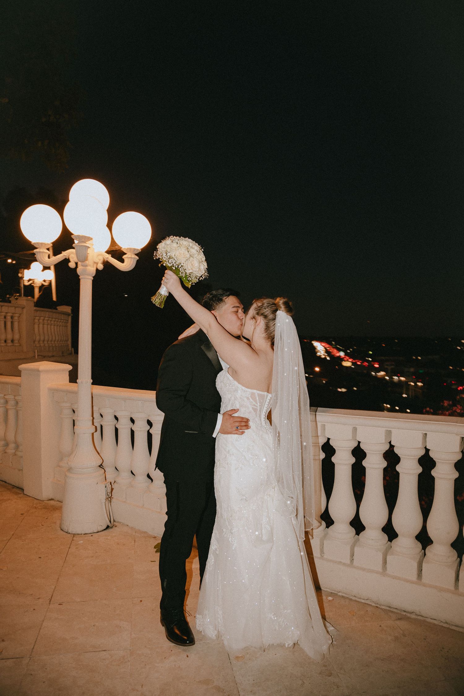 Wedding couple shares an intimate moment at night on a balcony with decorative lighting.