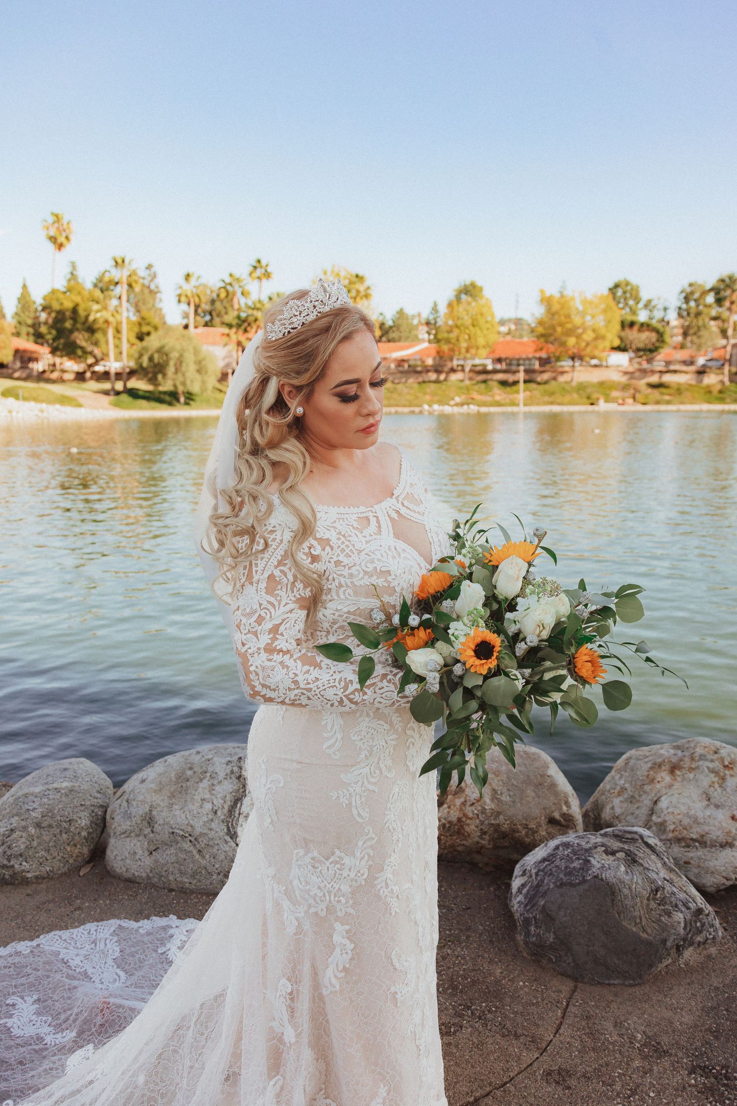 Bride in lace wedding gown holds sunflower bouquet by lakeside on sunny autumn day.