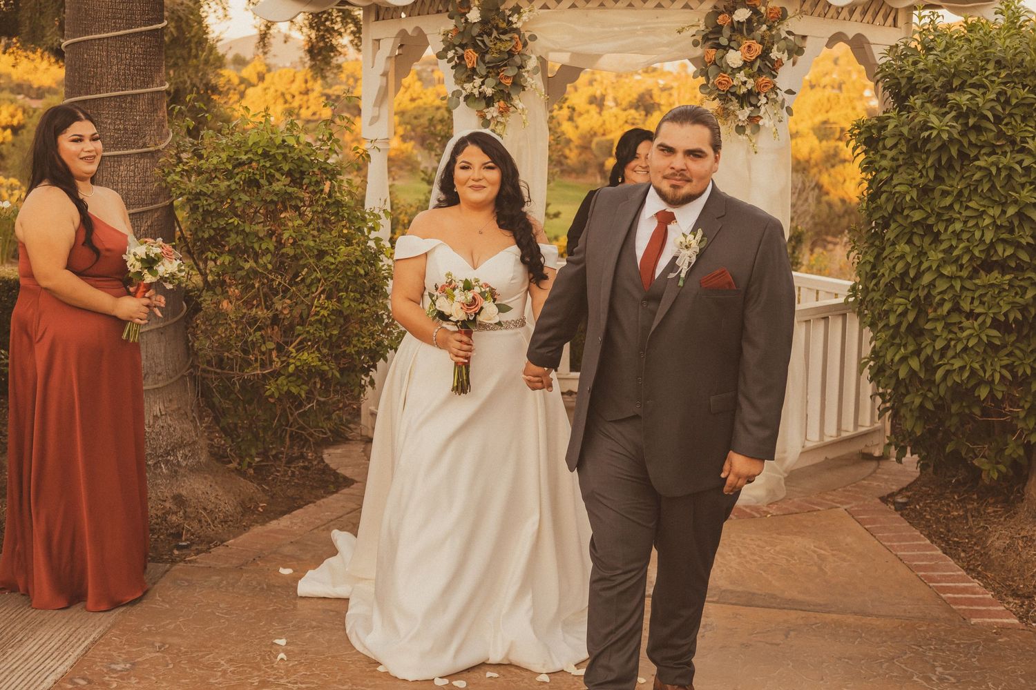 A wedding party walks down a path with a white gazebo and palm trees in the background.