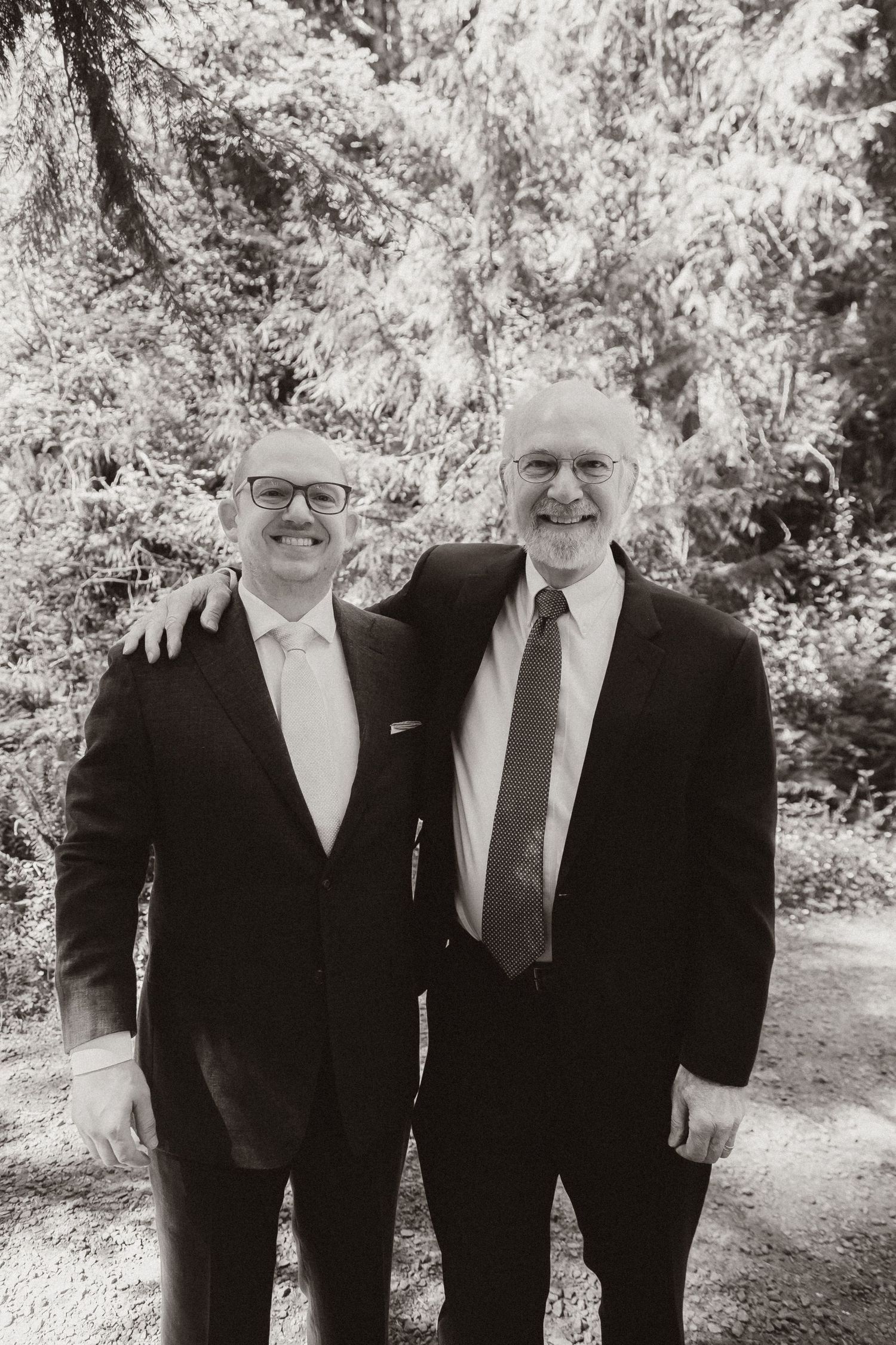 Two men in suits pose together for a formal black and white portrait outdoors with trees in the background.
