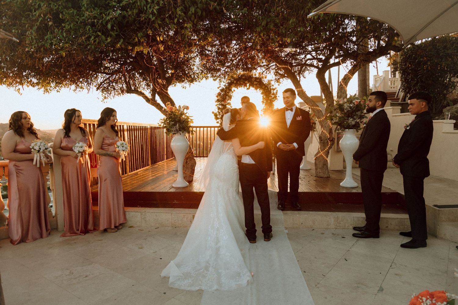 A golden sunset illuminates a wedding ceremony under trees with attendants in pink dresses.
