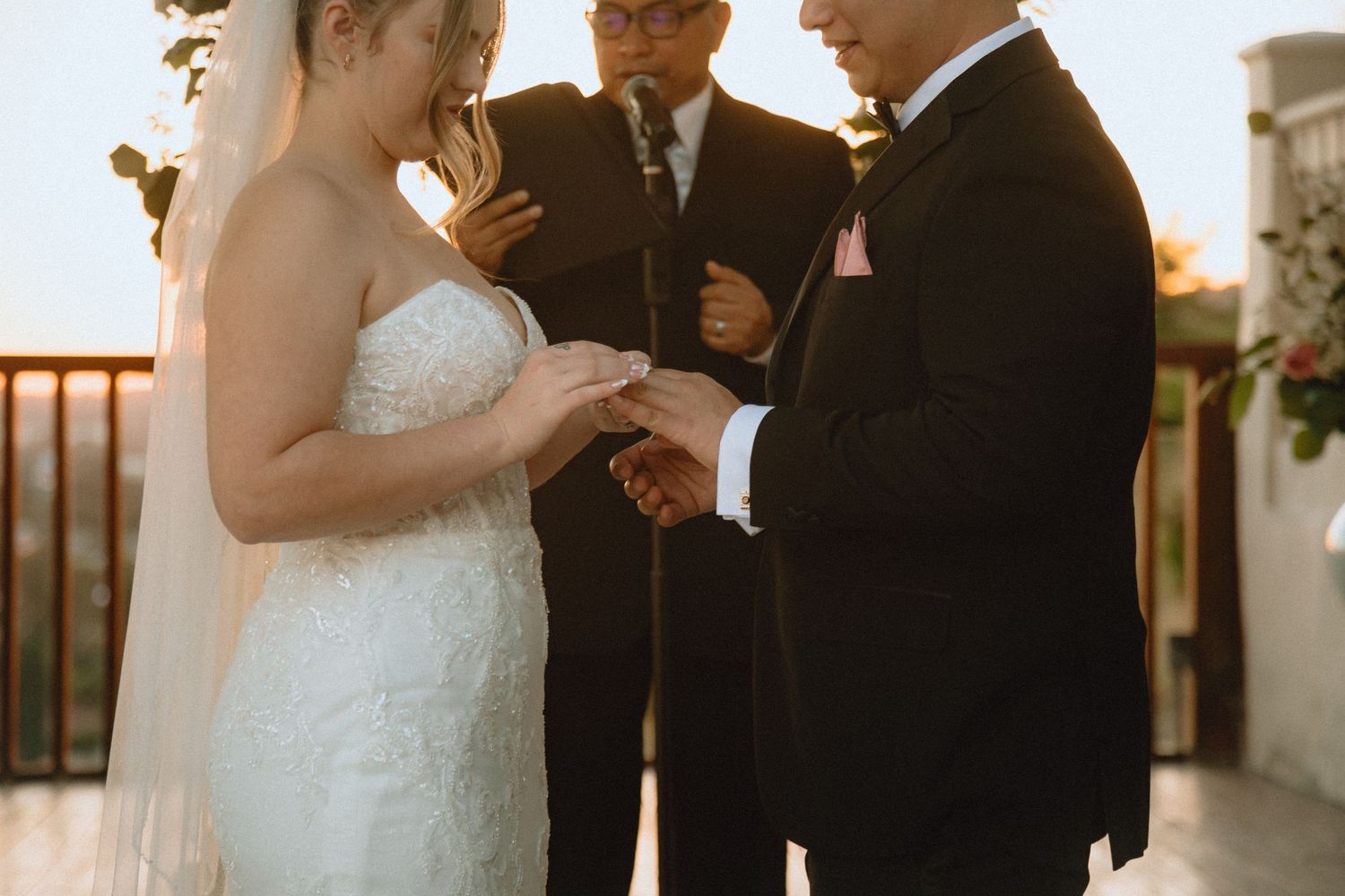 A couple exchanges wedding rings during their outdoor ceremony beneath string lights and floral decor.