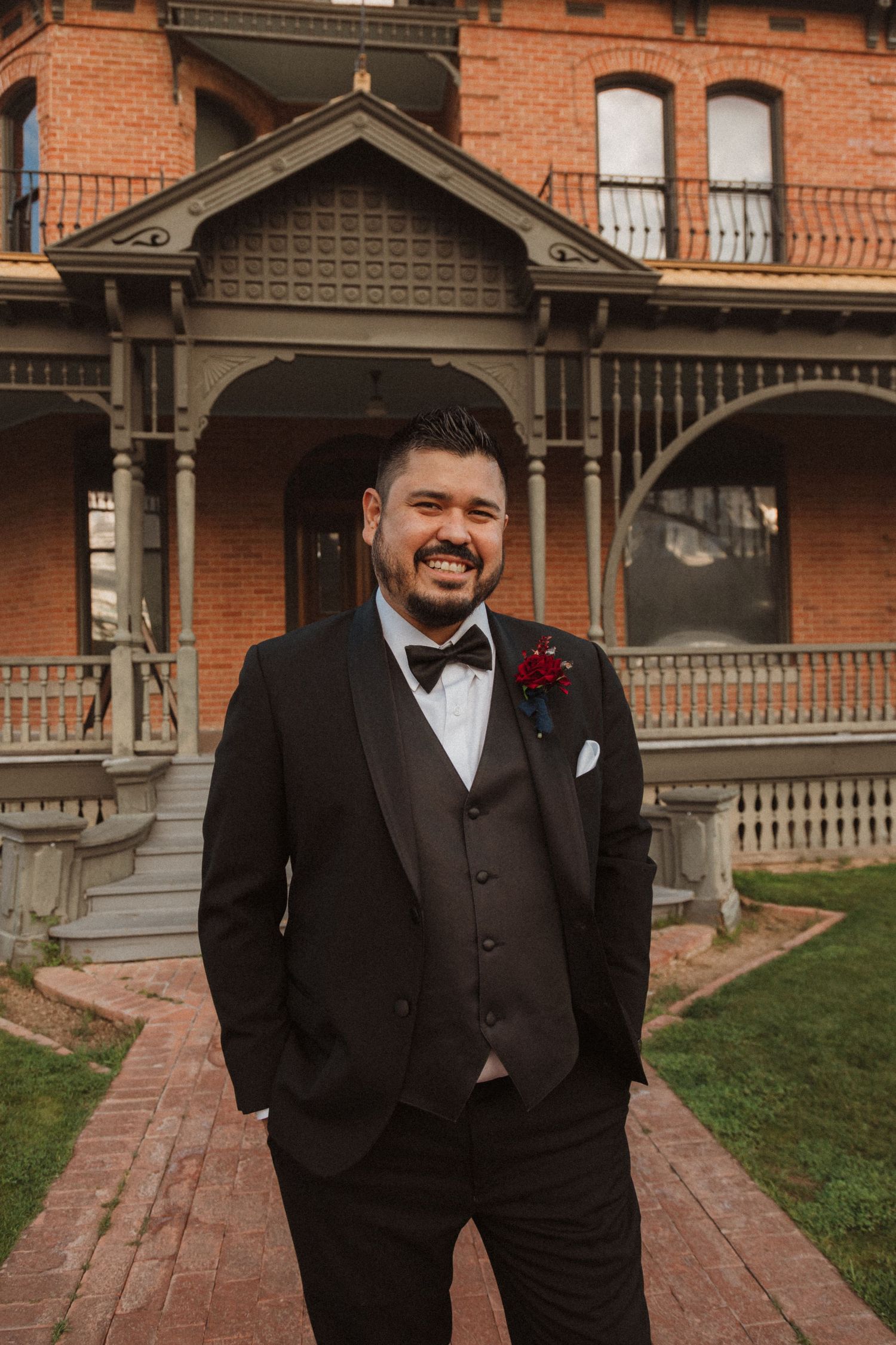 Smiling groom in formal attire stands before a historic red brick mansion with detailed woodwork.
