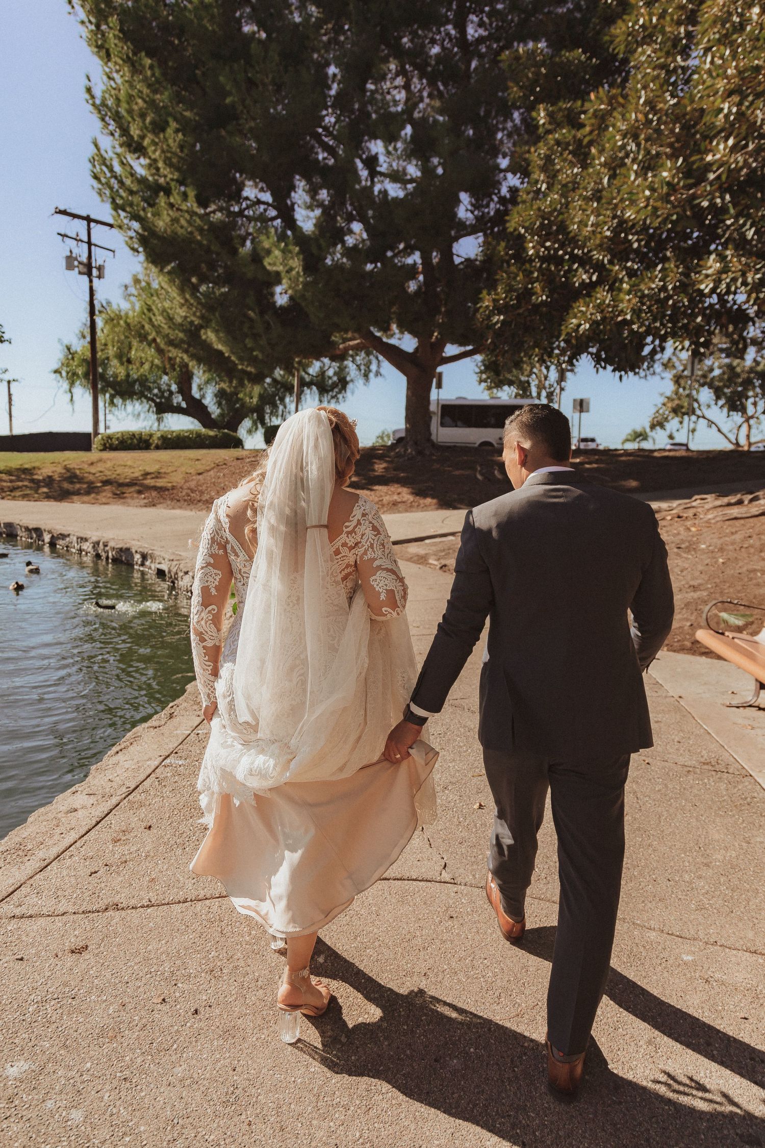 Newlyweds walking along waterfront path on sunny day with bride's dress trailing behind.