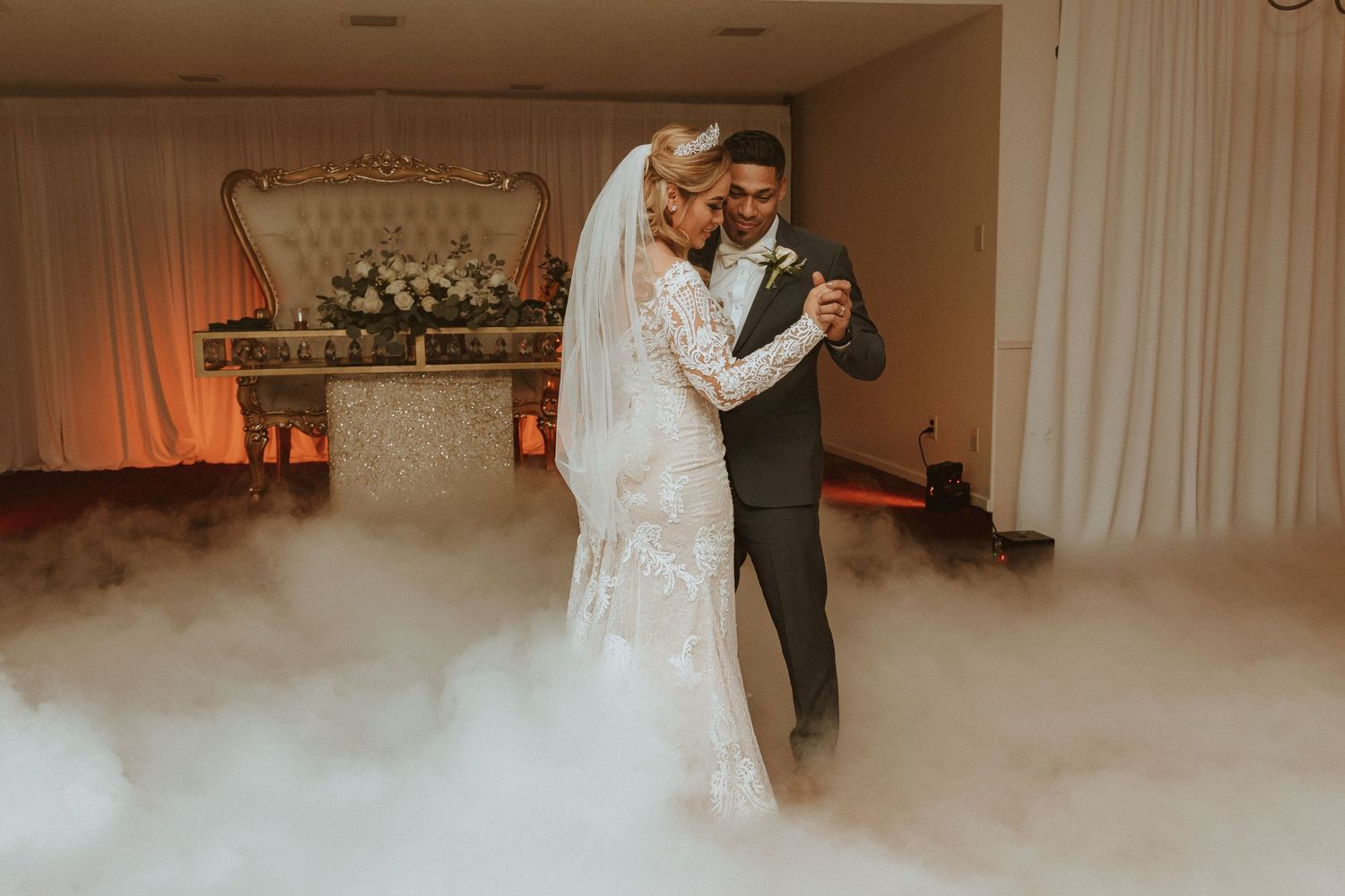 Romantic dance captured in elegant wedding venue with flowing fog and decorative floral backdrop.