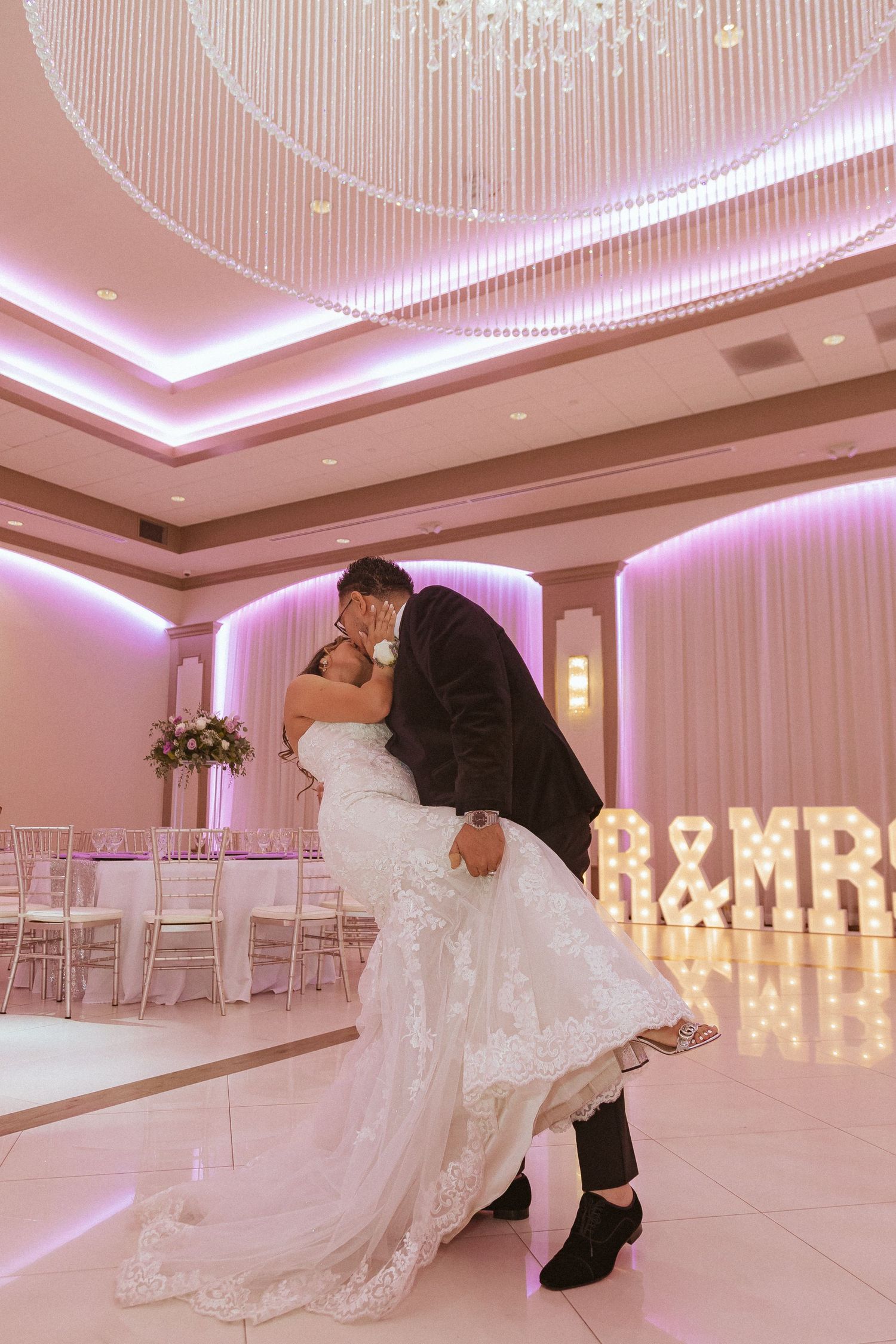 Wedding couple embraces in front of illuminated MR & MRS sign in an elegantly lit reception venue.