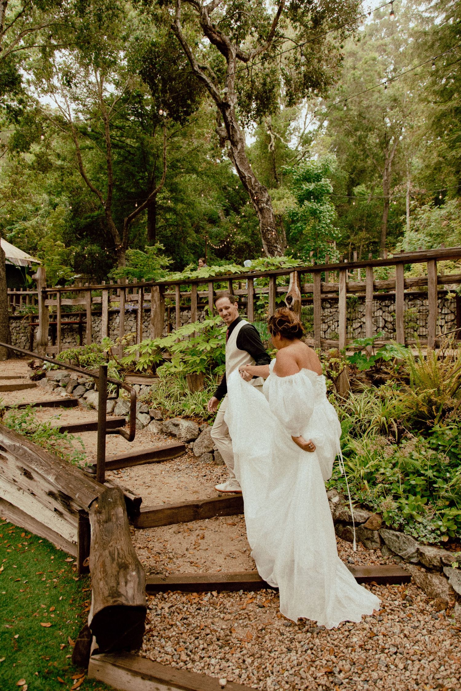 Two people in wedding attire ascend rustic wooden steps surrounded by lush garden landscaping.