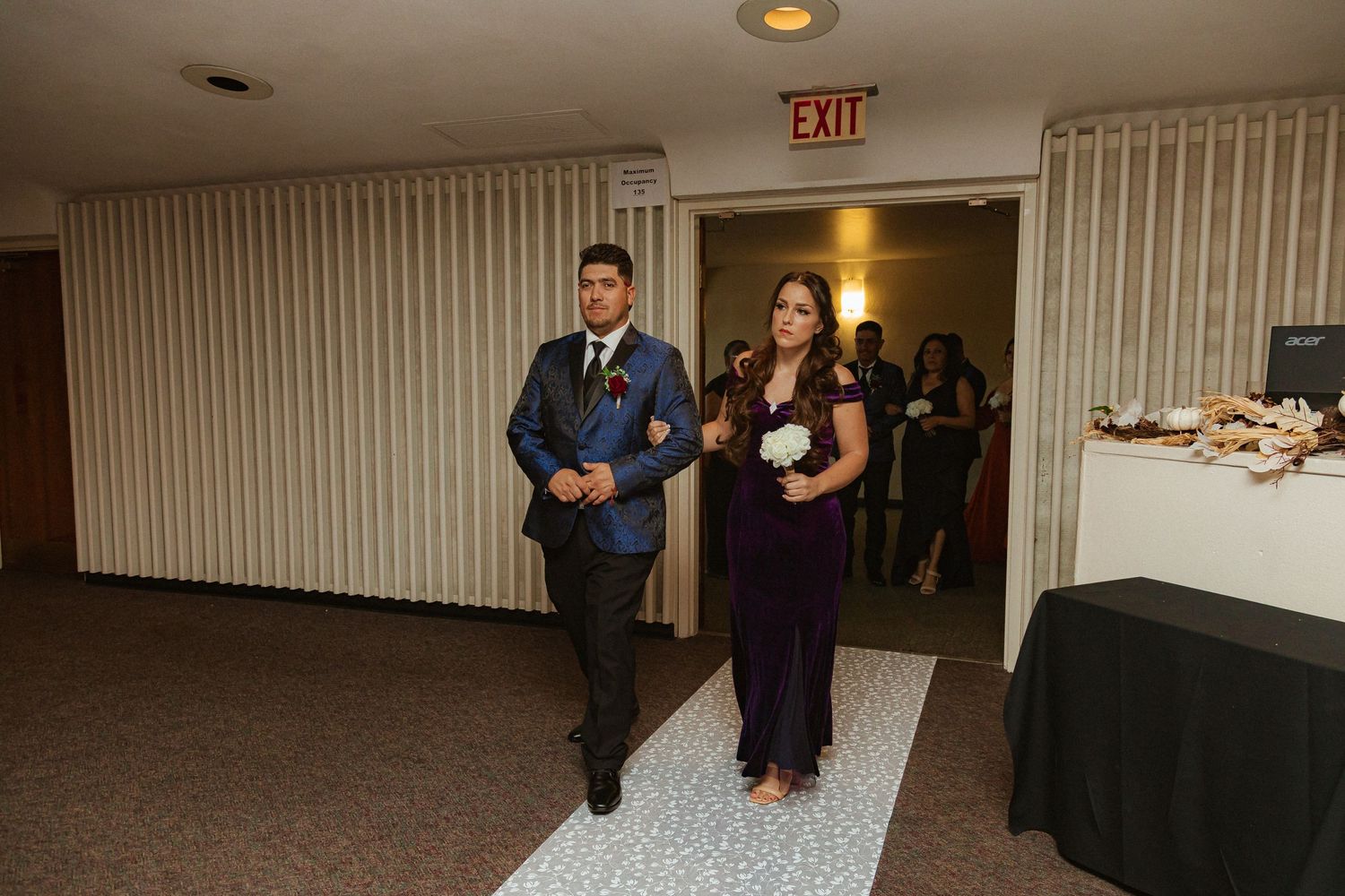 Wedding party members in formal attire walk down a carpeted hallway during reception entrance processional.