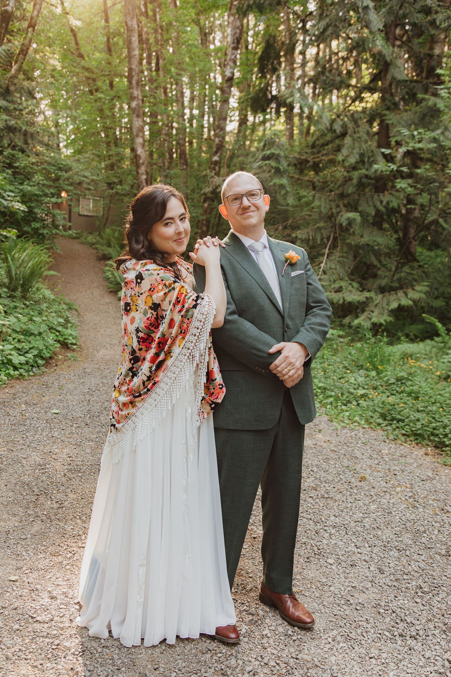 A wedding couple poses on a forest path, bride wearing a floral shawl over white dress.