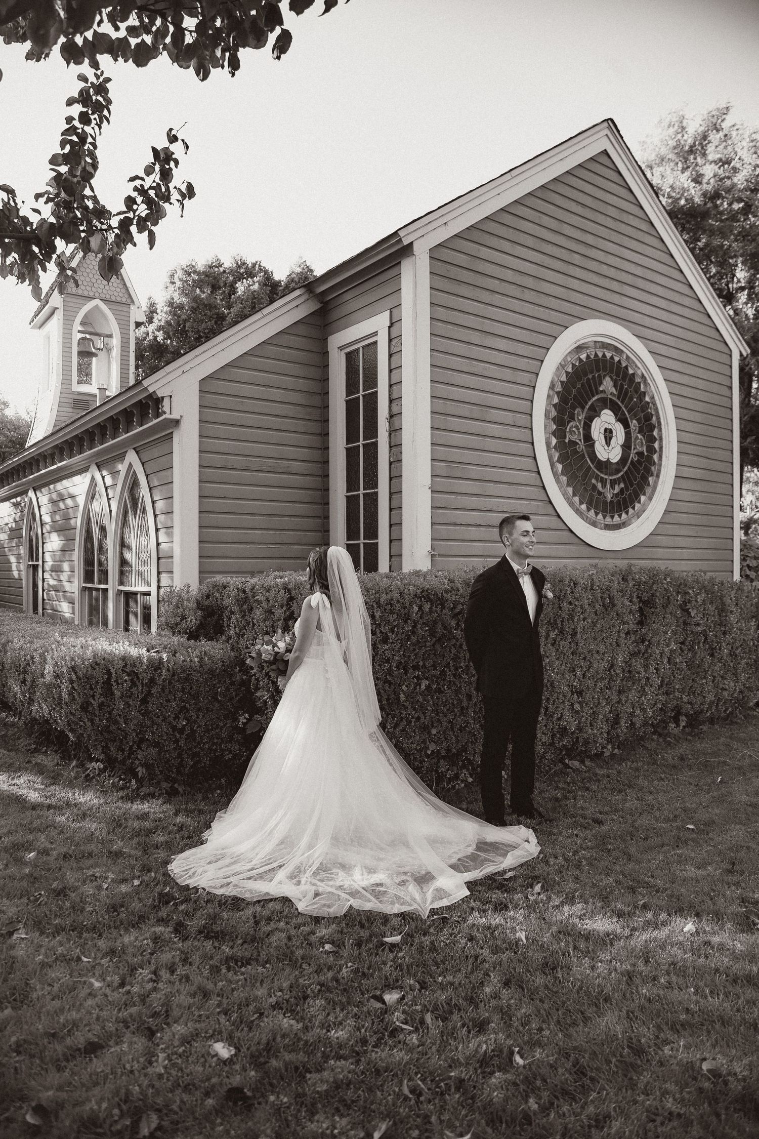 Black and white photo of couple in wedding attire outside chapel with long train dress.