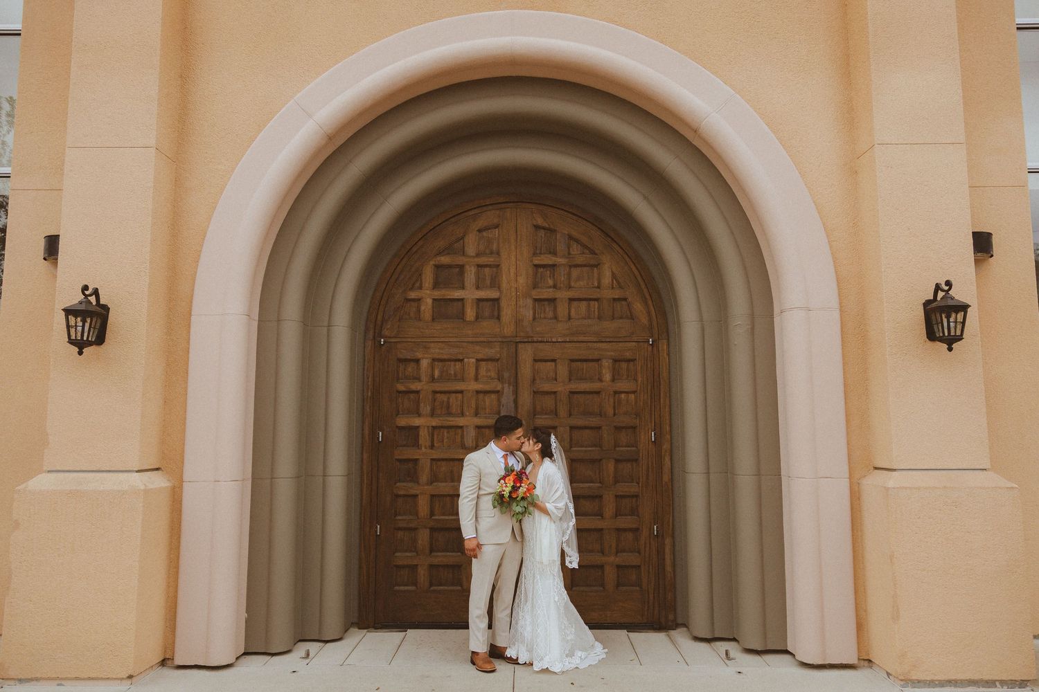 Intimate wedding portrait against ornate wooden church doors and peach-colored arched entrance.