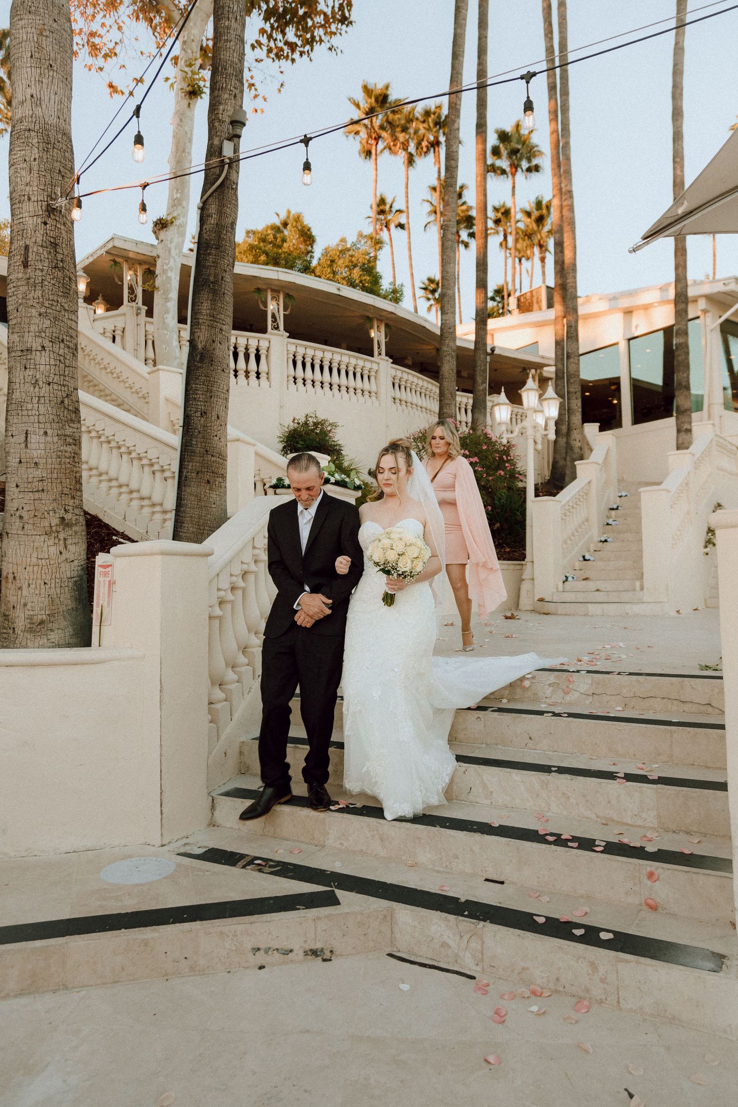 An outdoor wedding scene on grand white stairs with hanging lights and palm trees creating a romantic atmosphere.