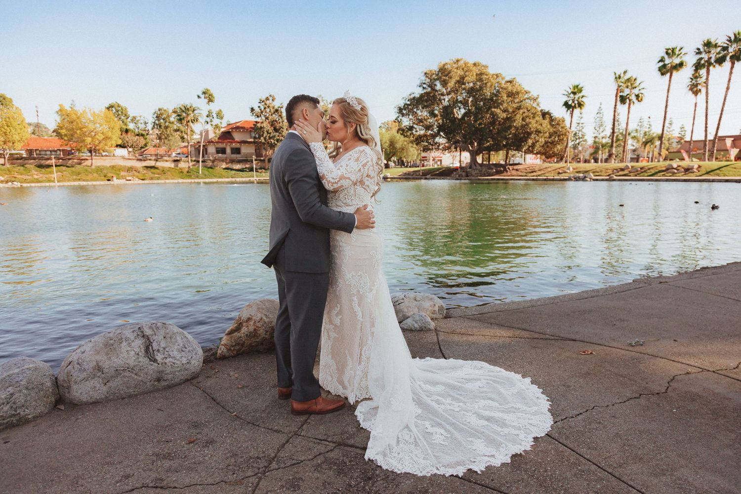 Bride in lace gown with long train poses with groom on lakeside path.