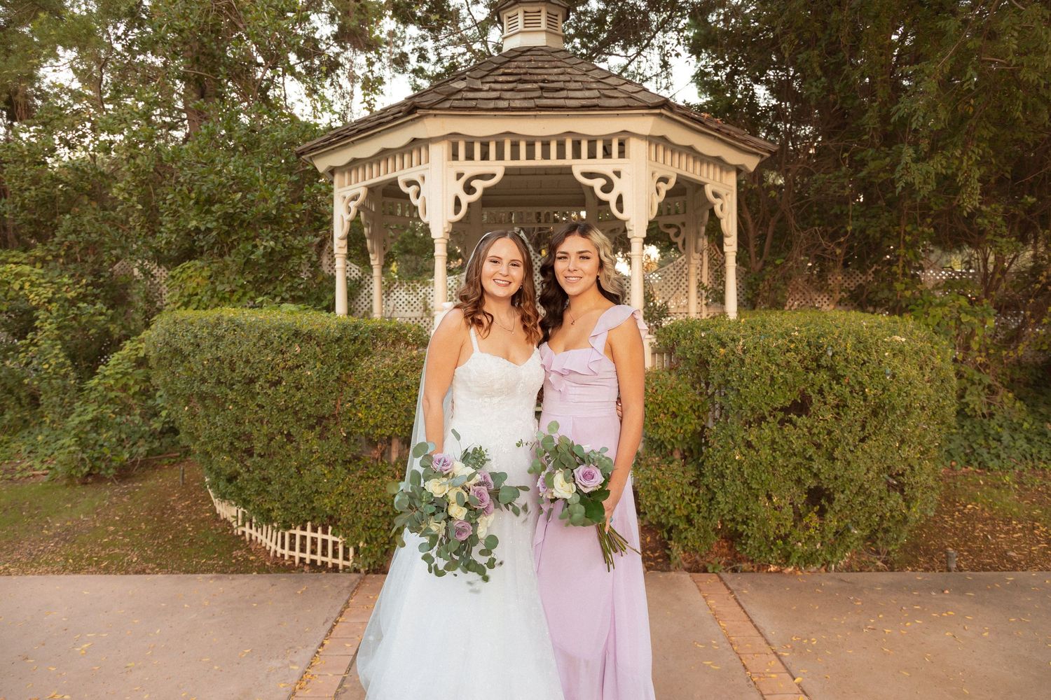 Two people in formal attire pose in front of a garden gazebo.