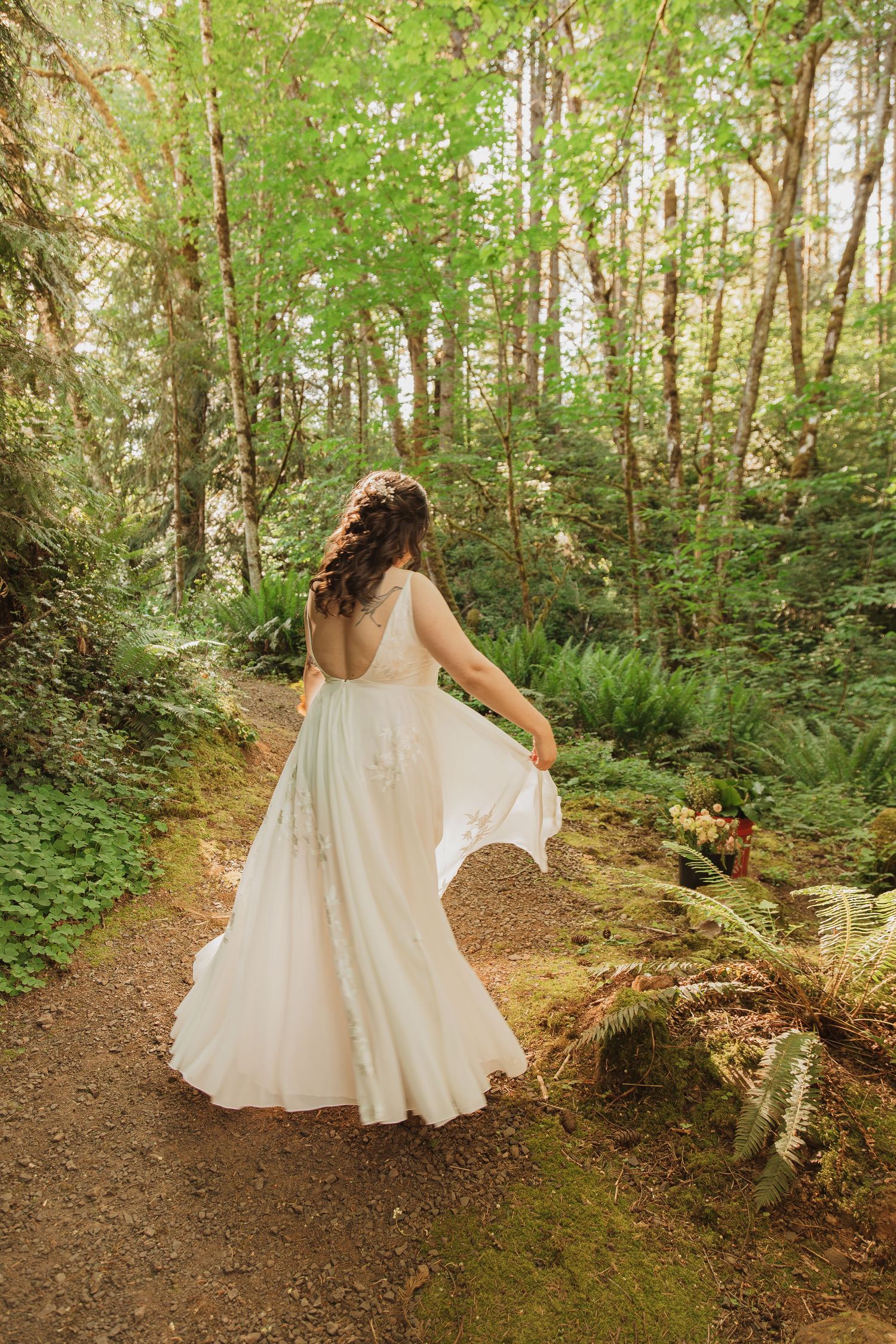 A person in a white wedding gown spins gracefully on a wooded trail surrounded by ferns and trees.