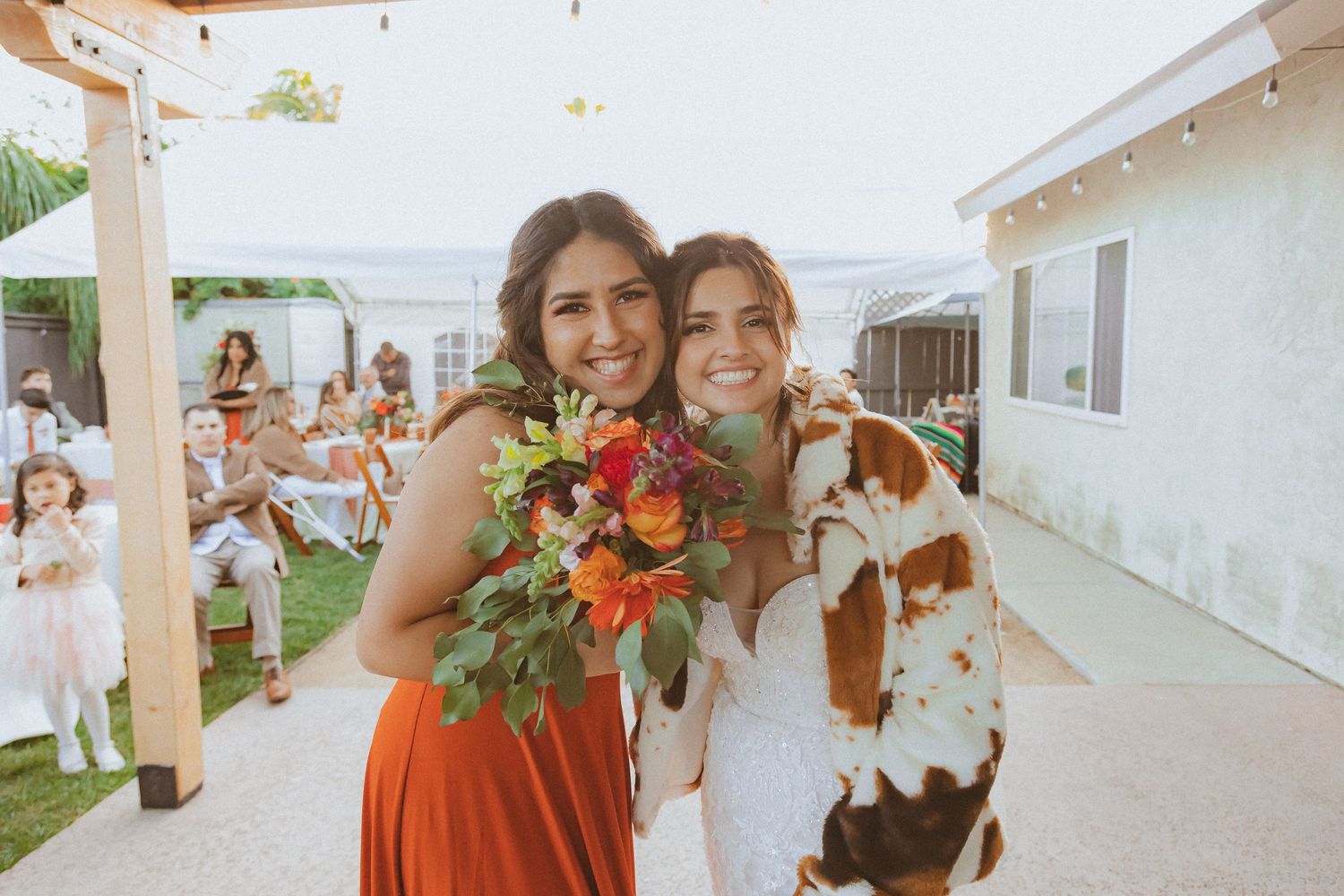 A bridesmaid in orange and bride embrace while holding colorful floral bouquets.