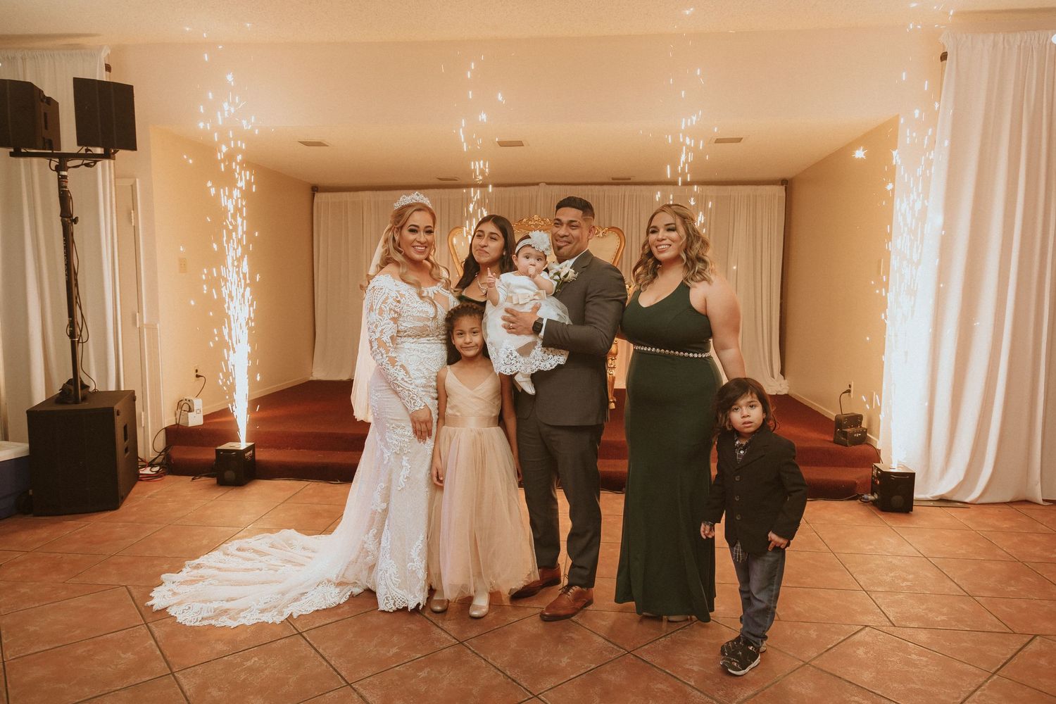 A joyful wedding party poses together in formal attire with sparkler fountains in the background.