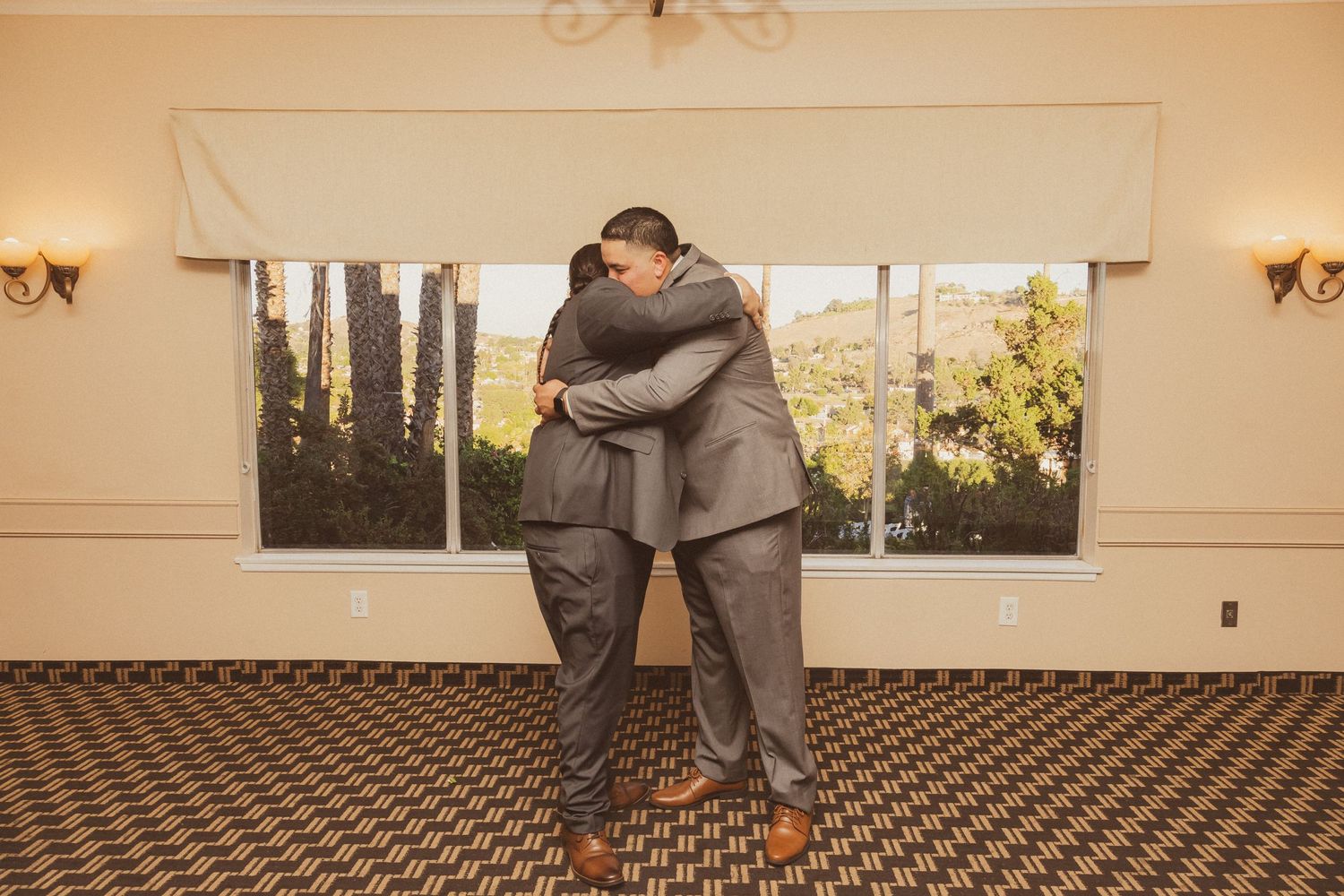 Two people in gray suits embracing in a room with patterned carpet and window view.
