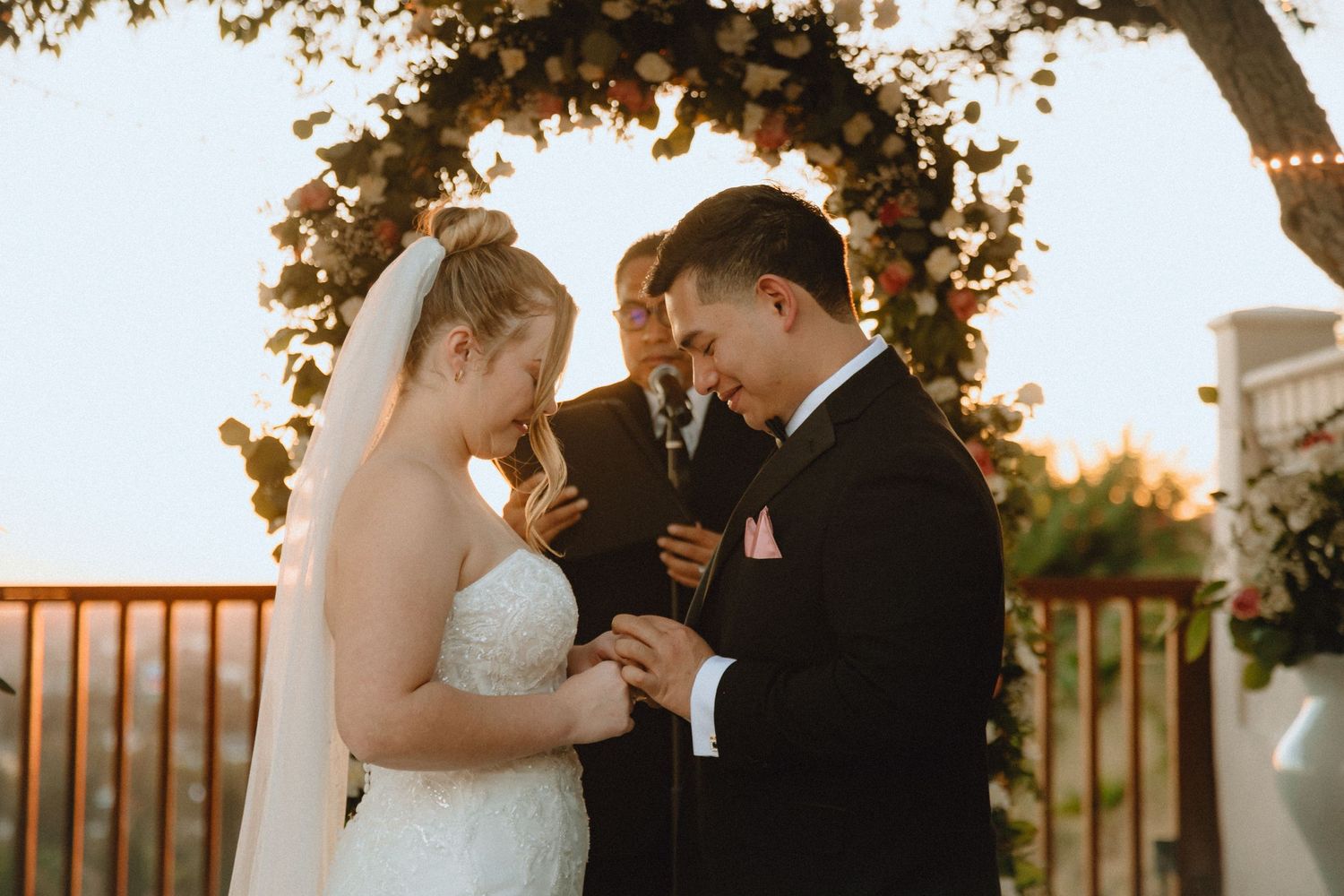 A wedding ceremony under a floral arch with string lights as the sun sets in the background.