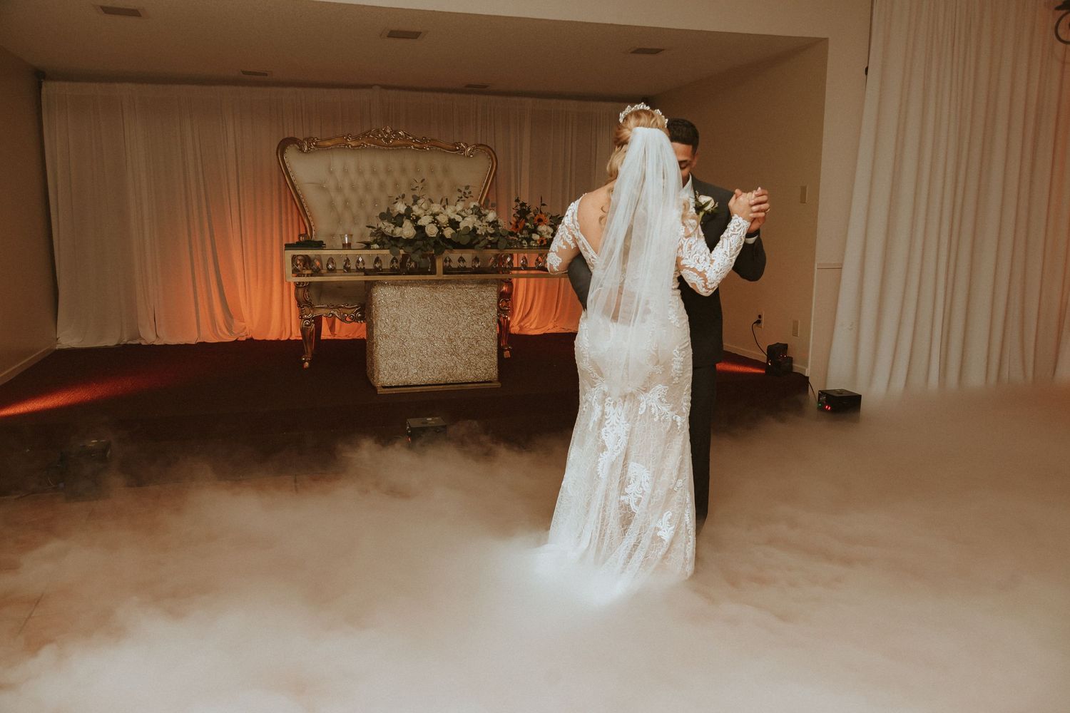 A bride in a lace gown stands amid atmospheric fog with floral decorations and soft lighting in the background.