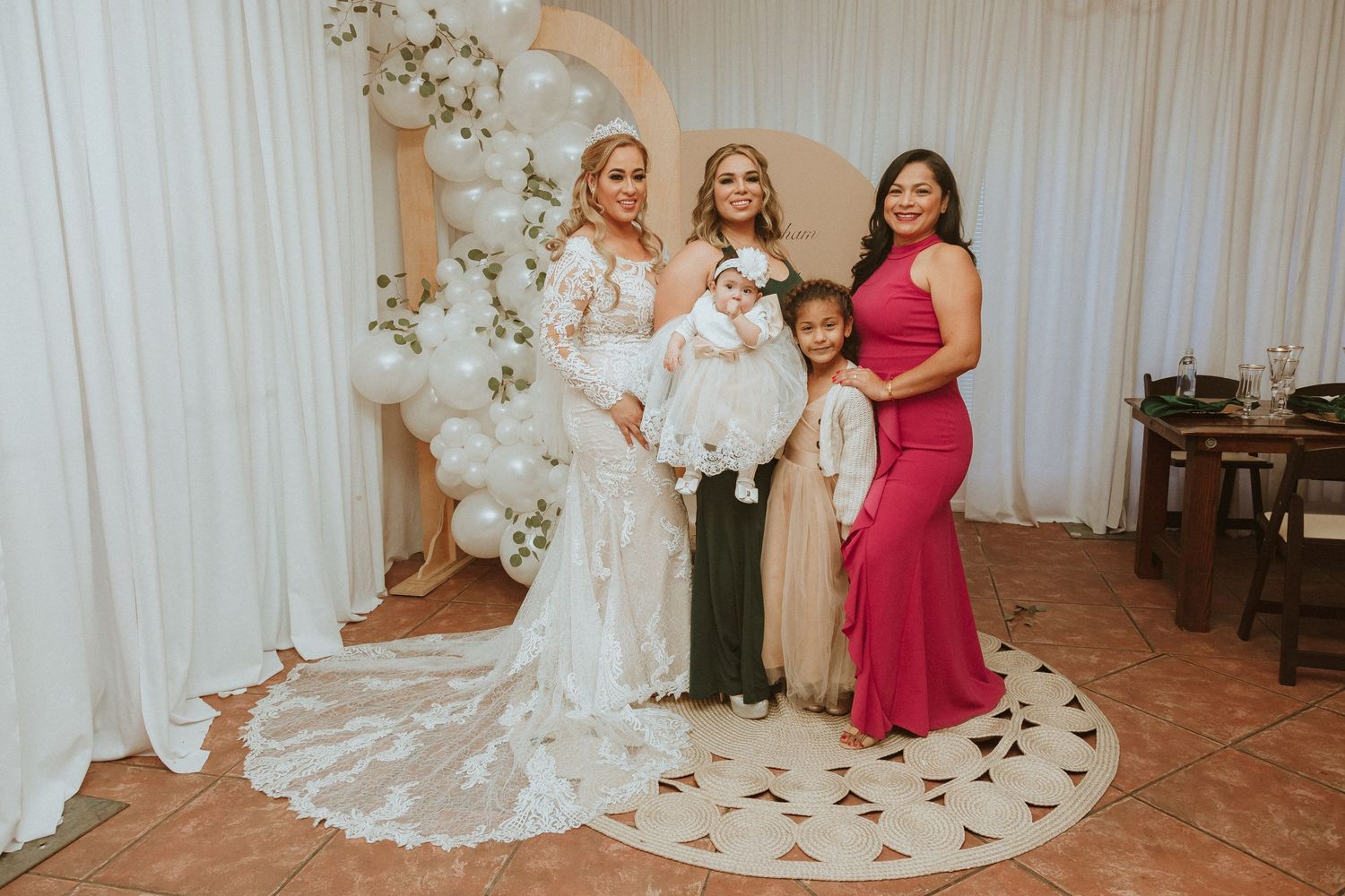 Wedding party poses against white floral arch backdrop with balloons and elegant gowns.