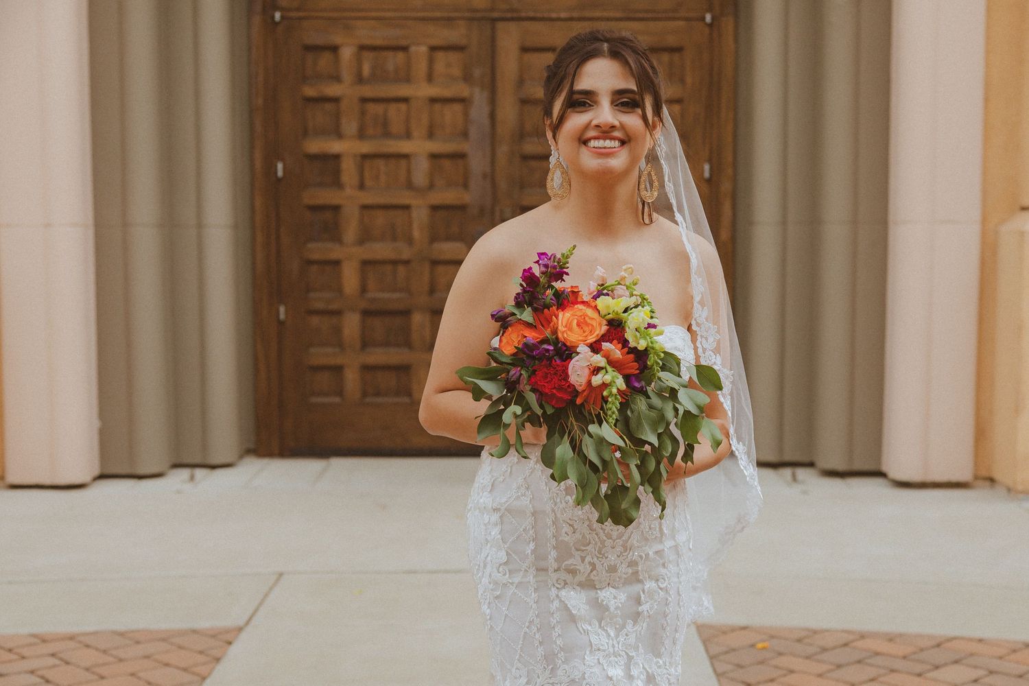 A happy bride in white lace gown holds colorful bouquet while standing before wooden church doors.