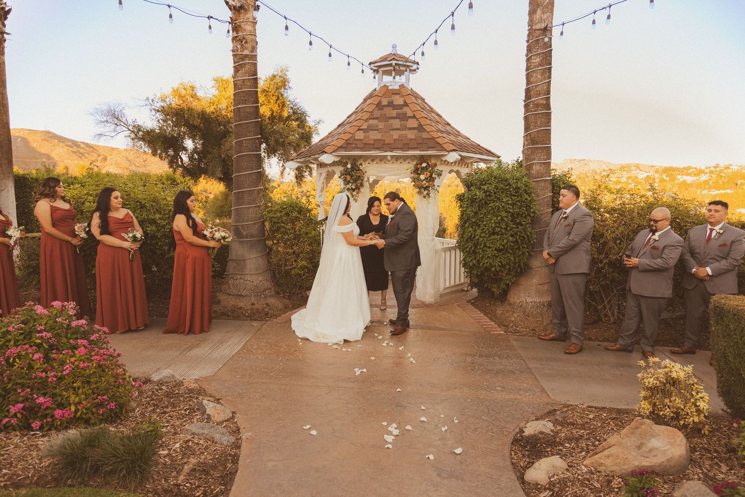 Wedding ceremony in progress at a rustic gazebo with burgundy-dressed bridesmaids and string lights.