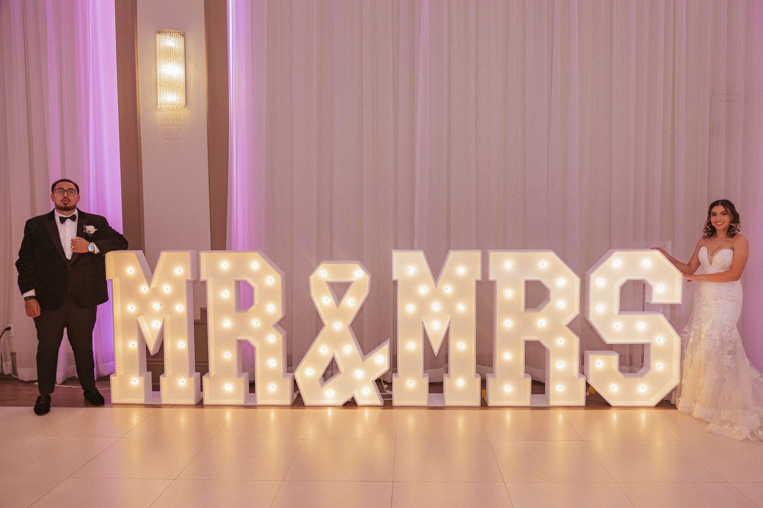 Wedding couple poses with illuminated MR & MRS marquee letters in reception hall.