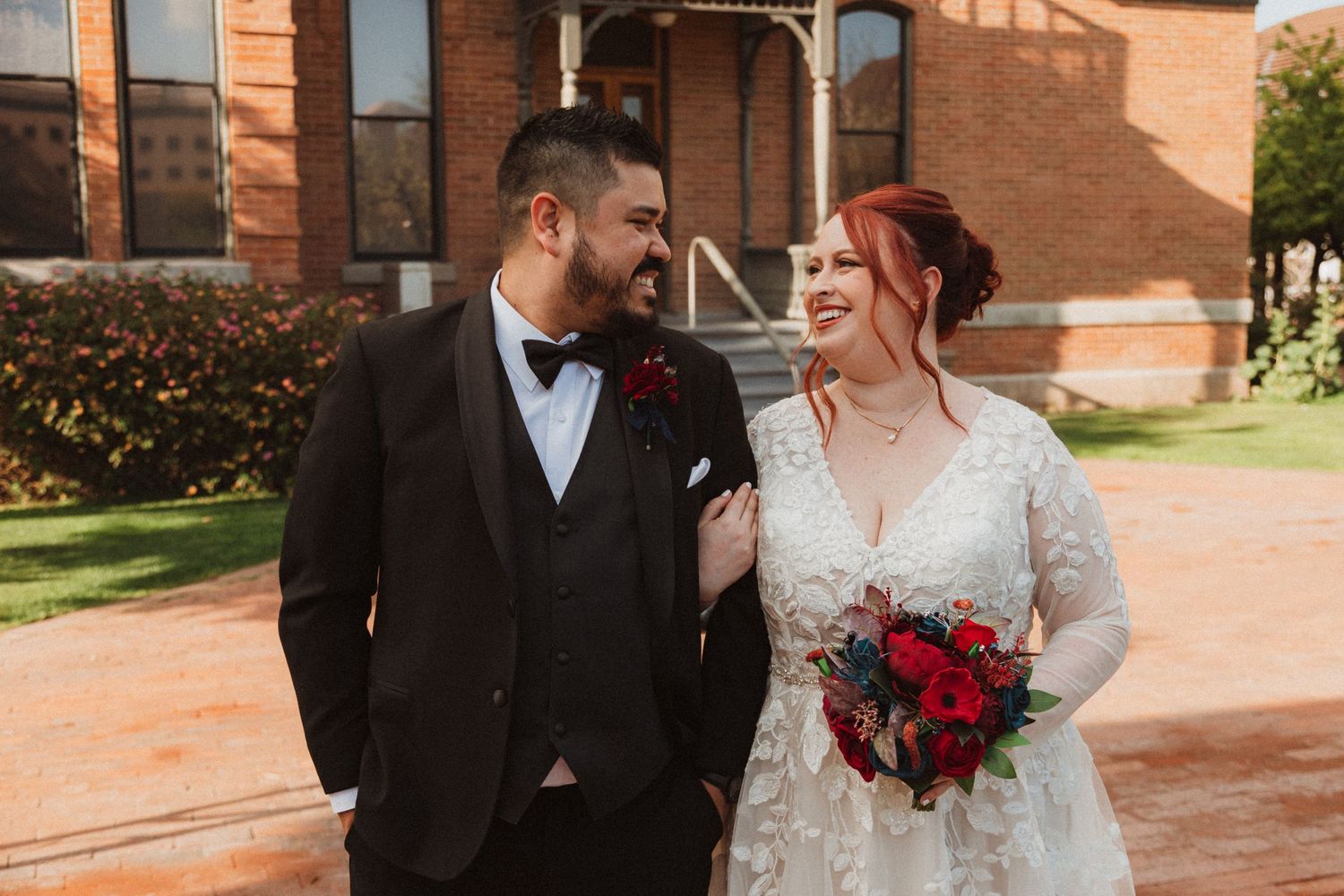 Couple in wedding attire standing outside historic brick building with red floral bouquet.