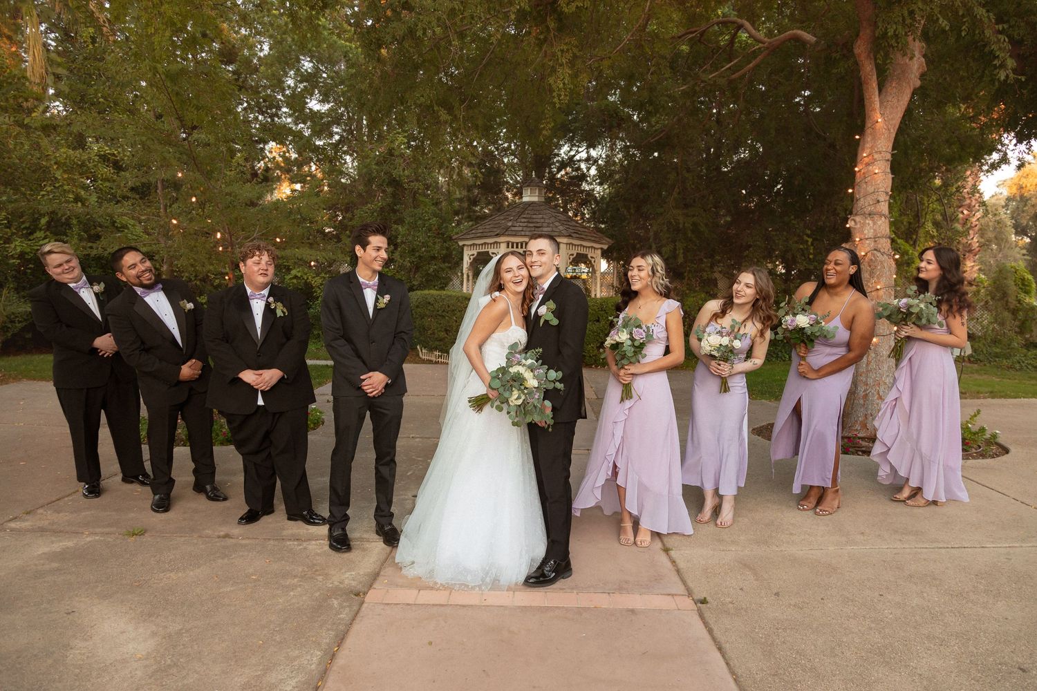 Wedding party lined up outdoors with bridesmaids wearing flowing lilac gowns and groomsmen in dark suits.