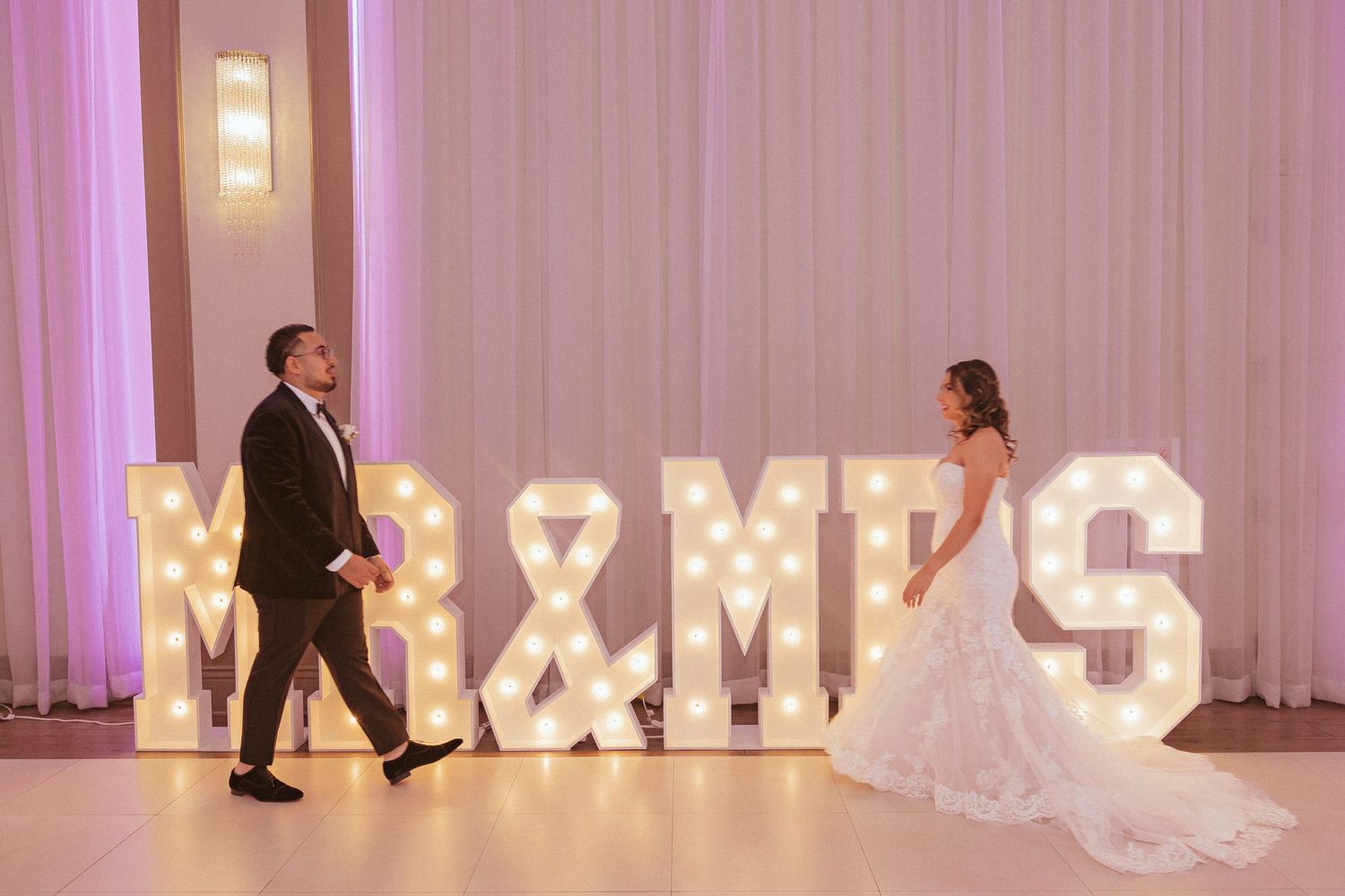 Couple walking towards illuminated MR & MRS marquee letters in pink-lit reception space.