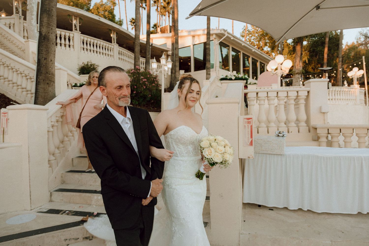 Elegant outdoor wedding scene with couple on white stairs surrounded by palm trees and decorative lighting.