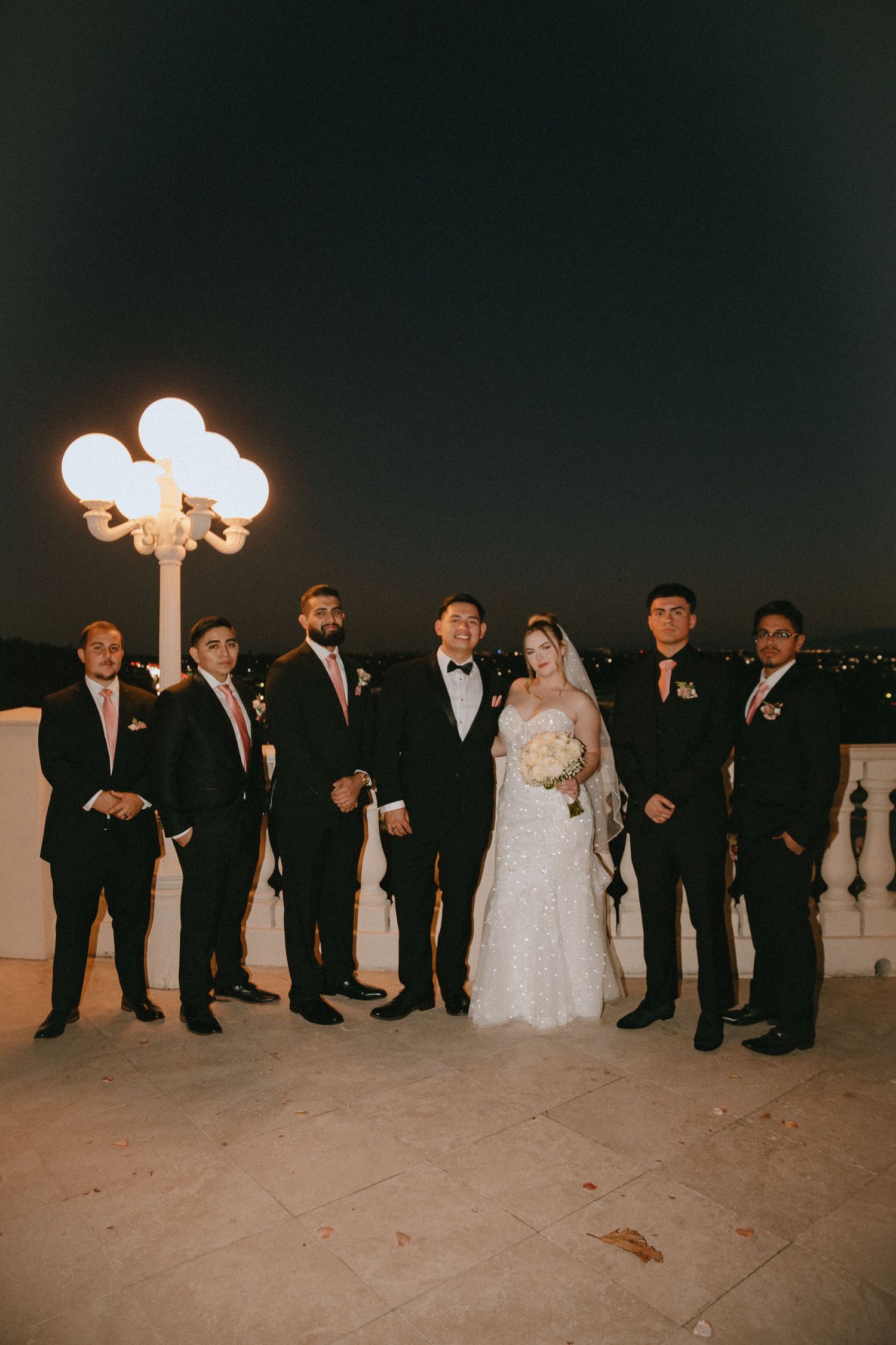 Wedding party stands elegantly on balcony at night with illuminated ornate lamp post in the background.