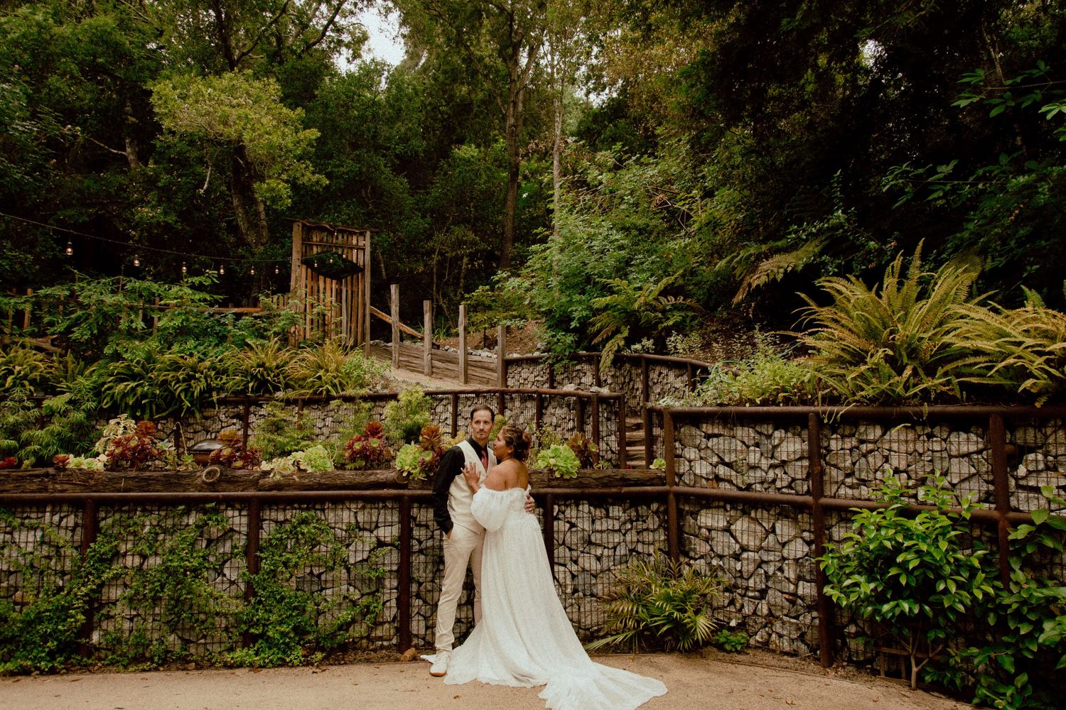Wedding couple embraces in front of a stone wall and lush garden terraces.