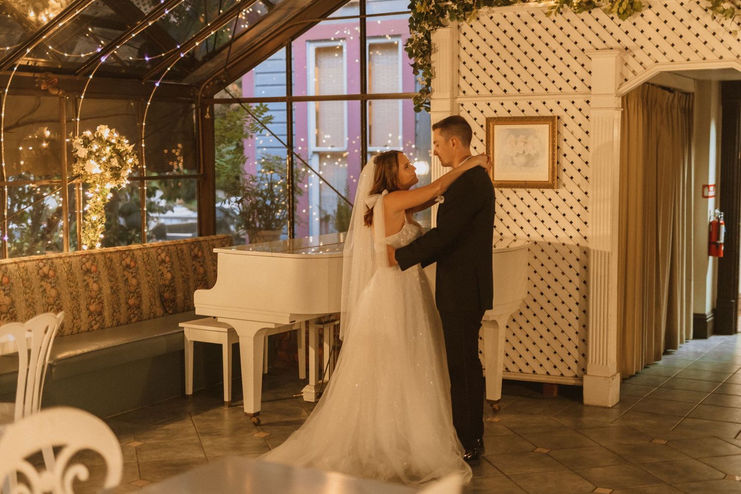 Wedding couple dances near white piano in venue with glass ceiling and string lights.