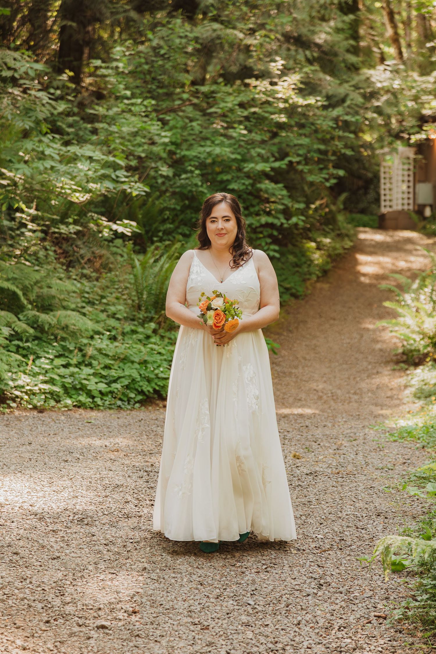 A person in a flowing white dress holds a bouquet while standing on a wooded trail.