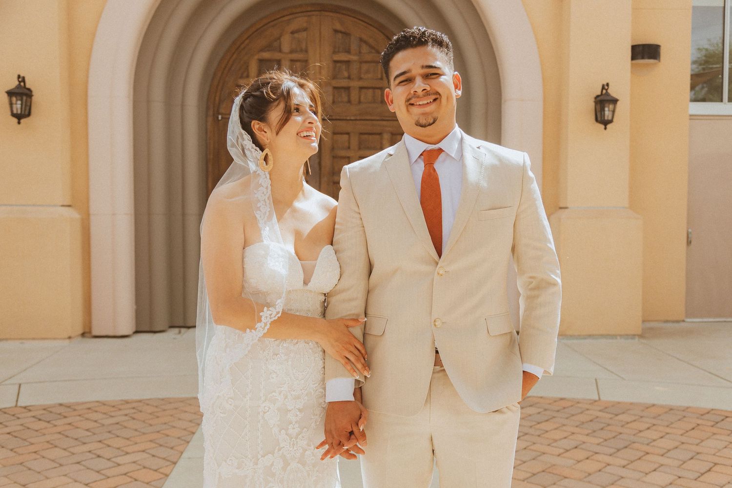 Newlyweds pose together in beige suit and lace wedding dress against Mediterranean-style architecture.