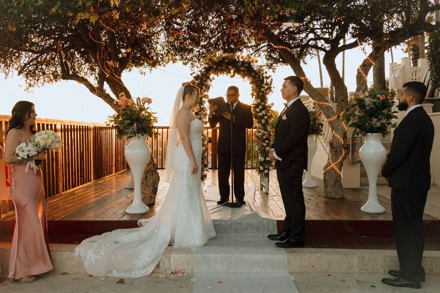 Golden hour wedding ceremony with bridesmaids in blush dresses and white floral decorations.