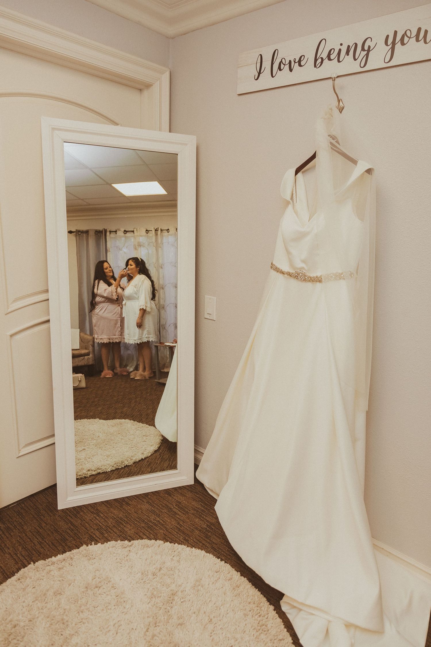 Wedding dress hangs beside full-length mirror in preparation room with 'I love being you' wall text.