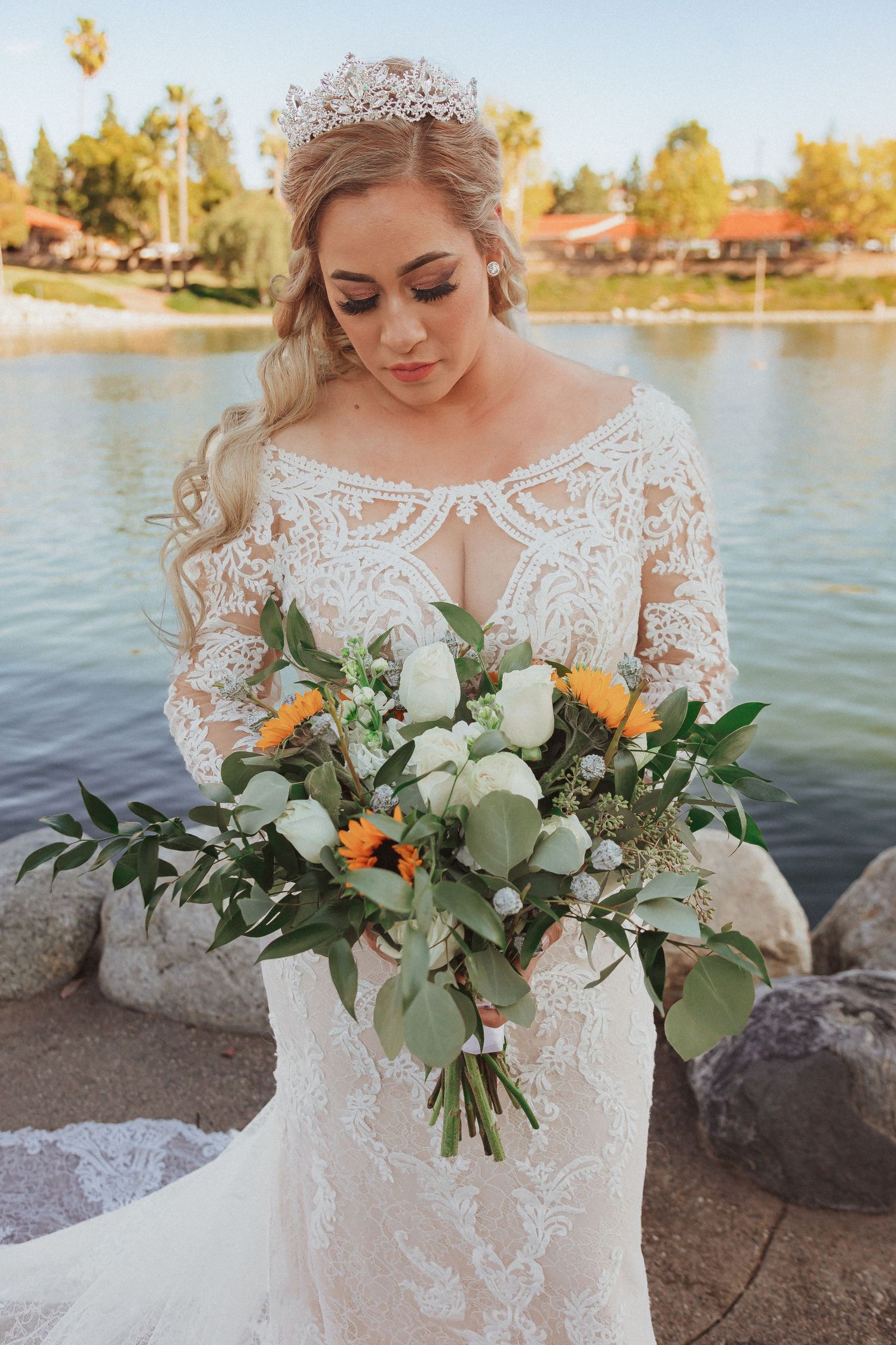 A bridal portrait by a lake featuring an elegant lace gown, delicate tiara, and natural bouquet with white and orange flowers.