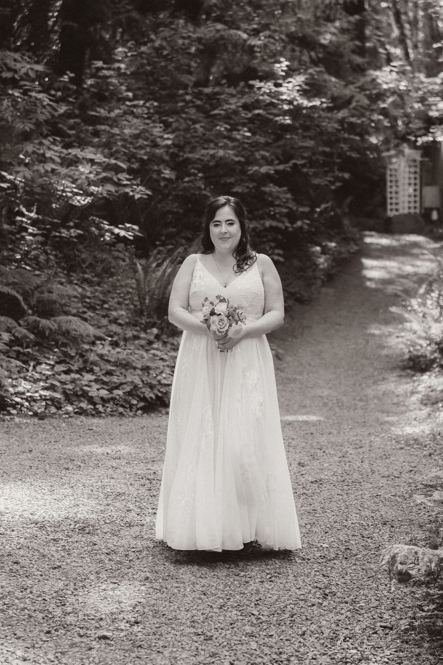 A person in a white wedding gown stands holding a bouquet on a forest path.