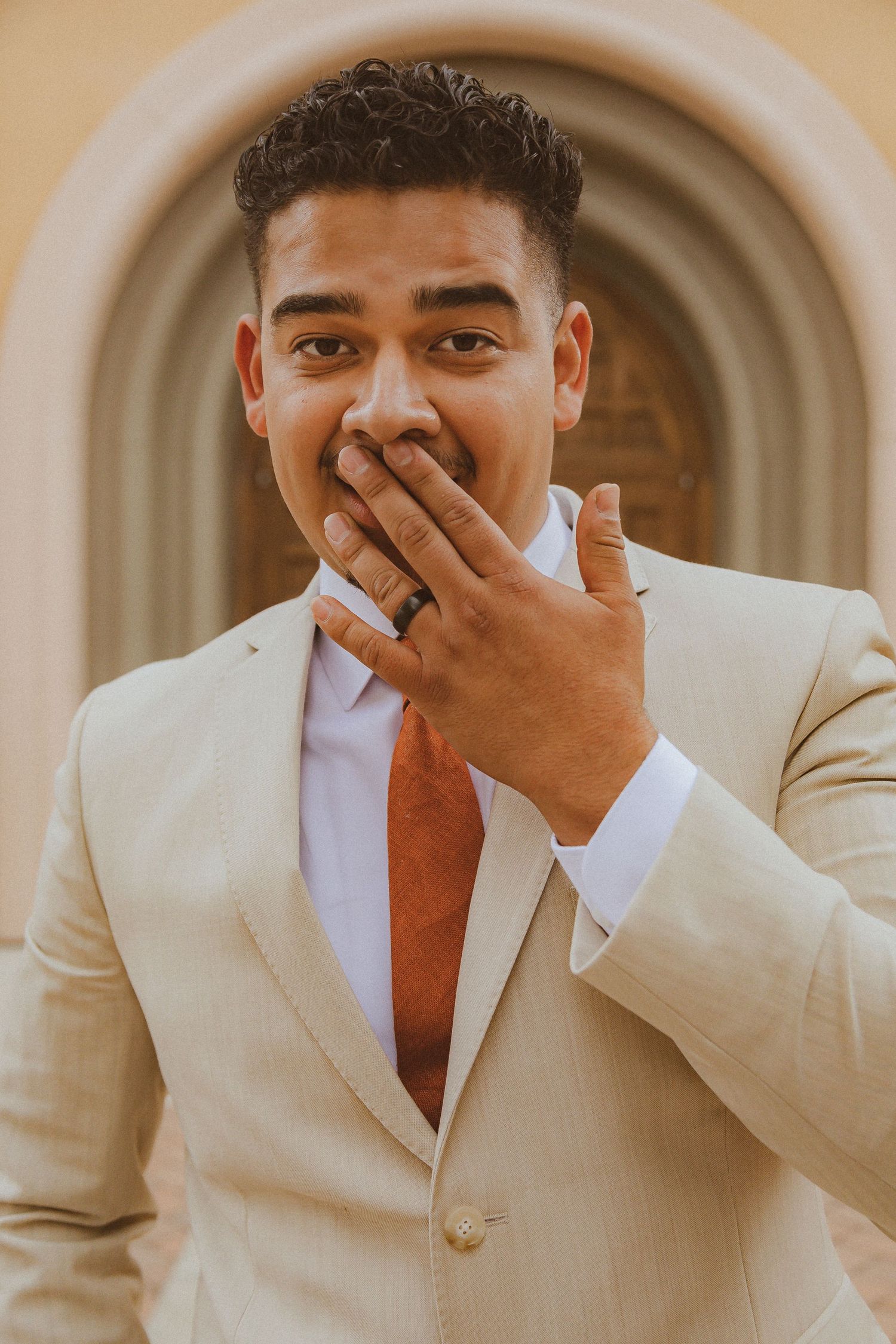 A groom in a beige suit and orange tie covers his mouth with excitement on his wedding day.