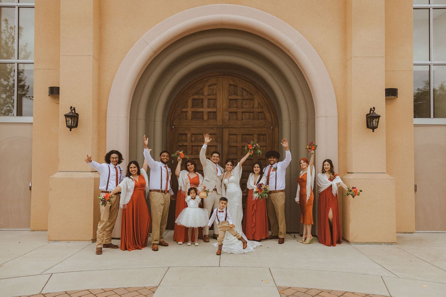 Energetic wedding party celebration photo with coral and khaki color scheme at church entrance.