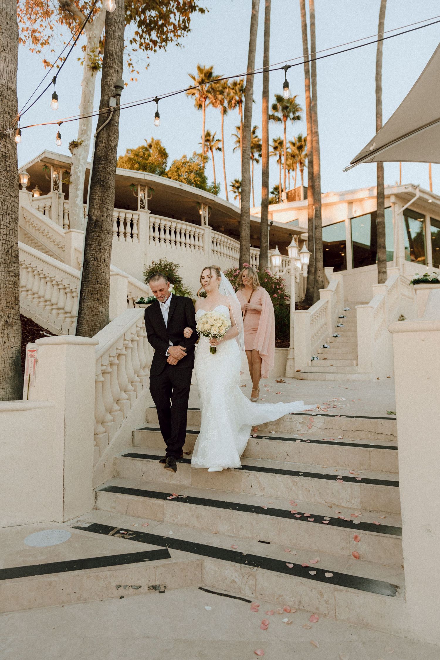 Wedding ceremony participants walking down white marble steps with palm trees and string lights in the background.