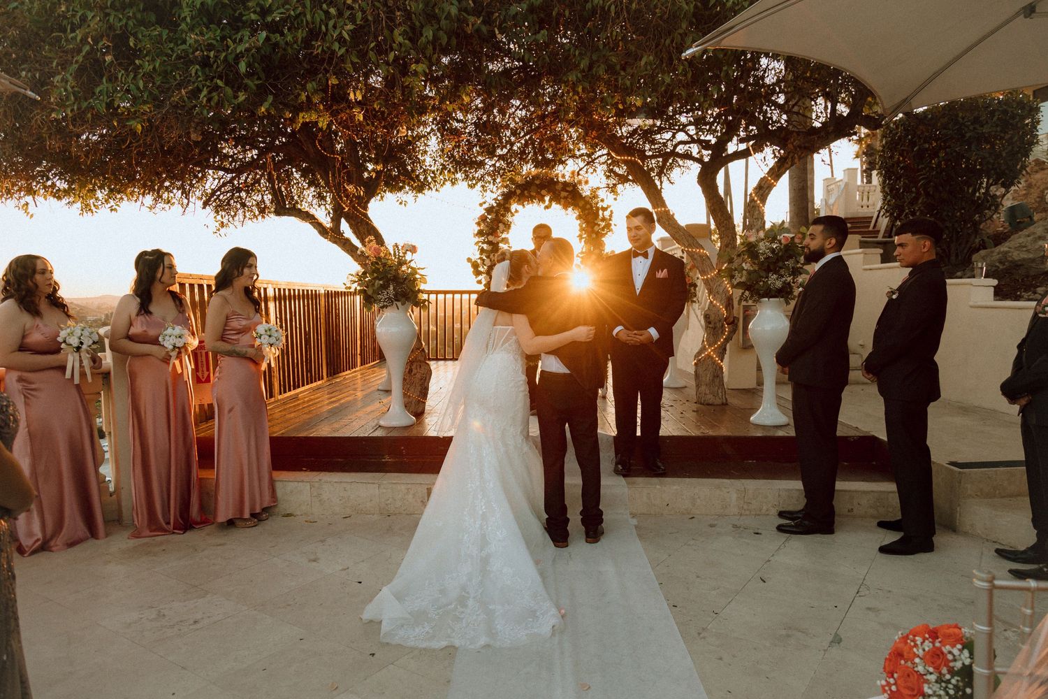 A sunset wedding ceremony with attendants in pink dresses under a tree canopy.