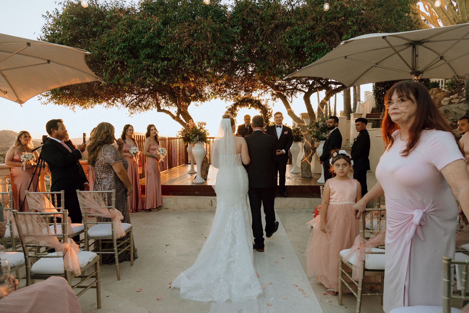 A bride in white lace walks down the aisle at an outdoor wedding ceremony.